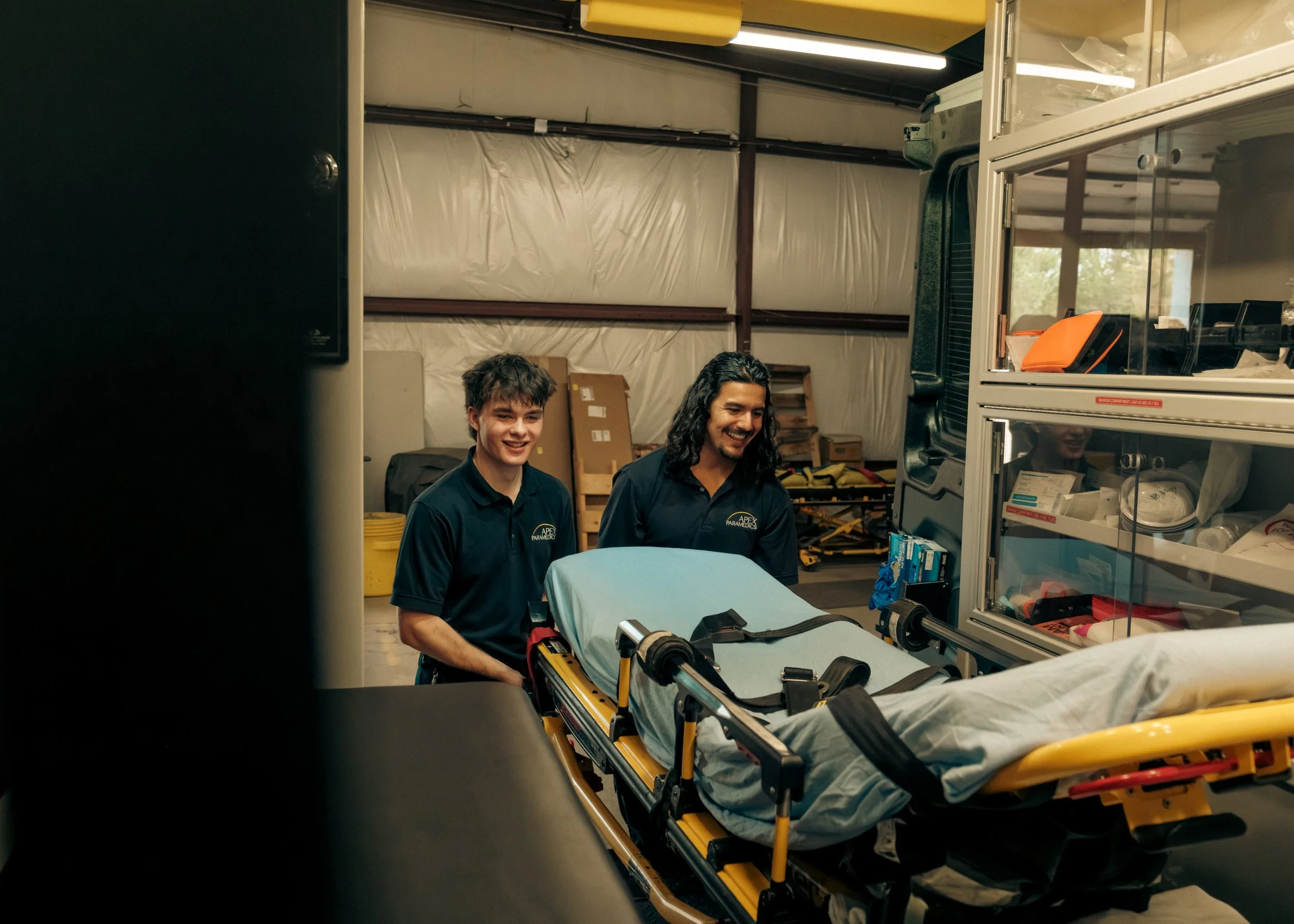 Two emergency medical personnel in navy uniforms with 'APEX PARAMEDICS' logo, standing beside a stretcher with a patient covered in a light blue sheet inside a storeroom or medical supply area with shelves and various medical supplies.