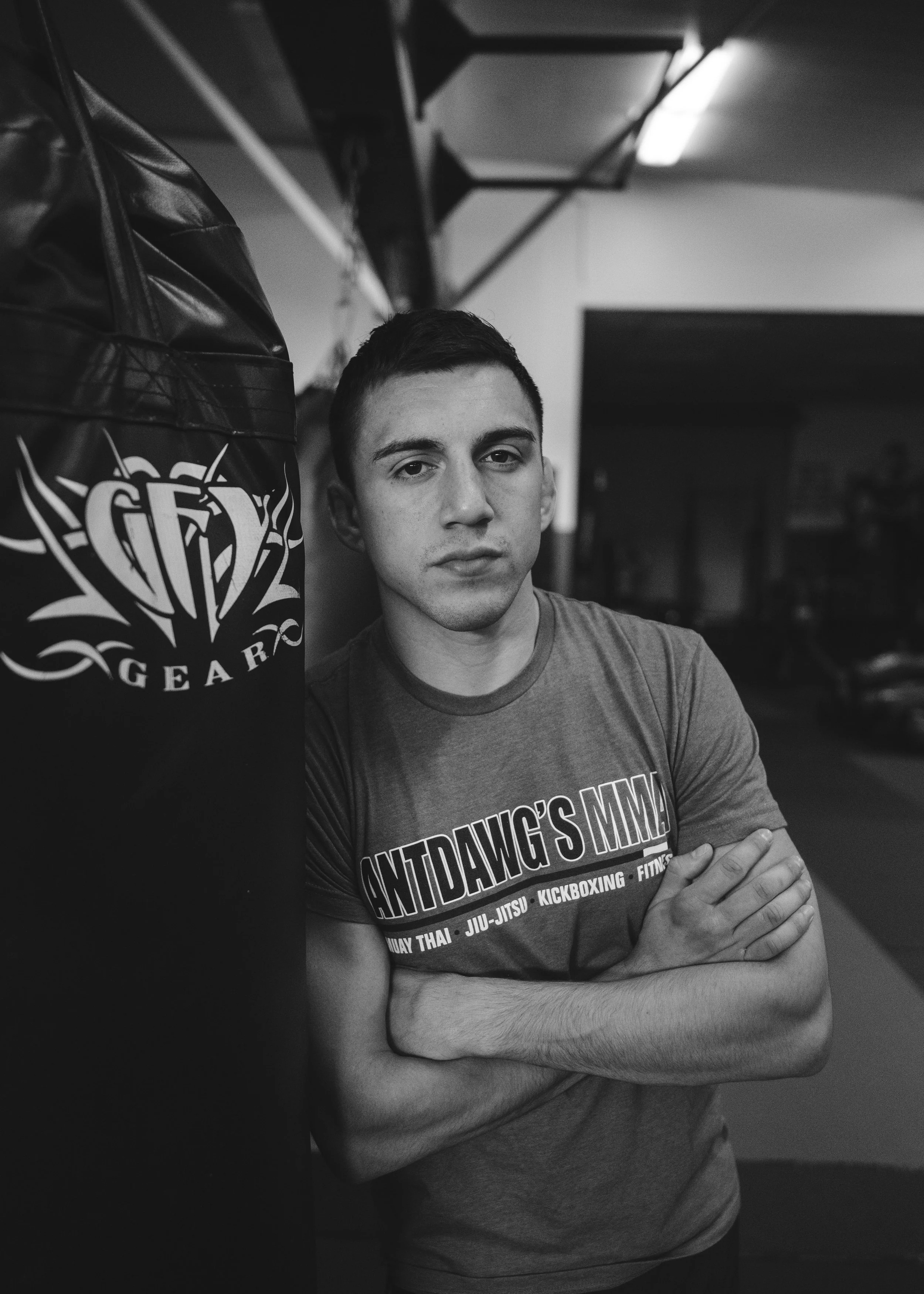 A young man with short dark hair and light skin is standing in front of a dented punching bag with a logo on it. He is wearing a T-shirt with the words "HUNTDAWG'S MMA" and other martial arts disciplines listed. His arms are crossed, and he looks dir
