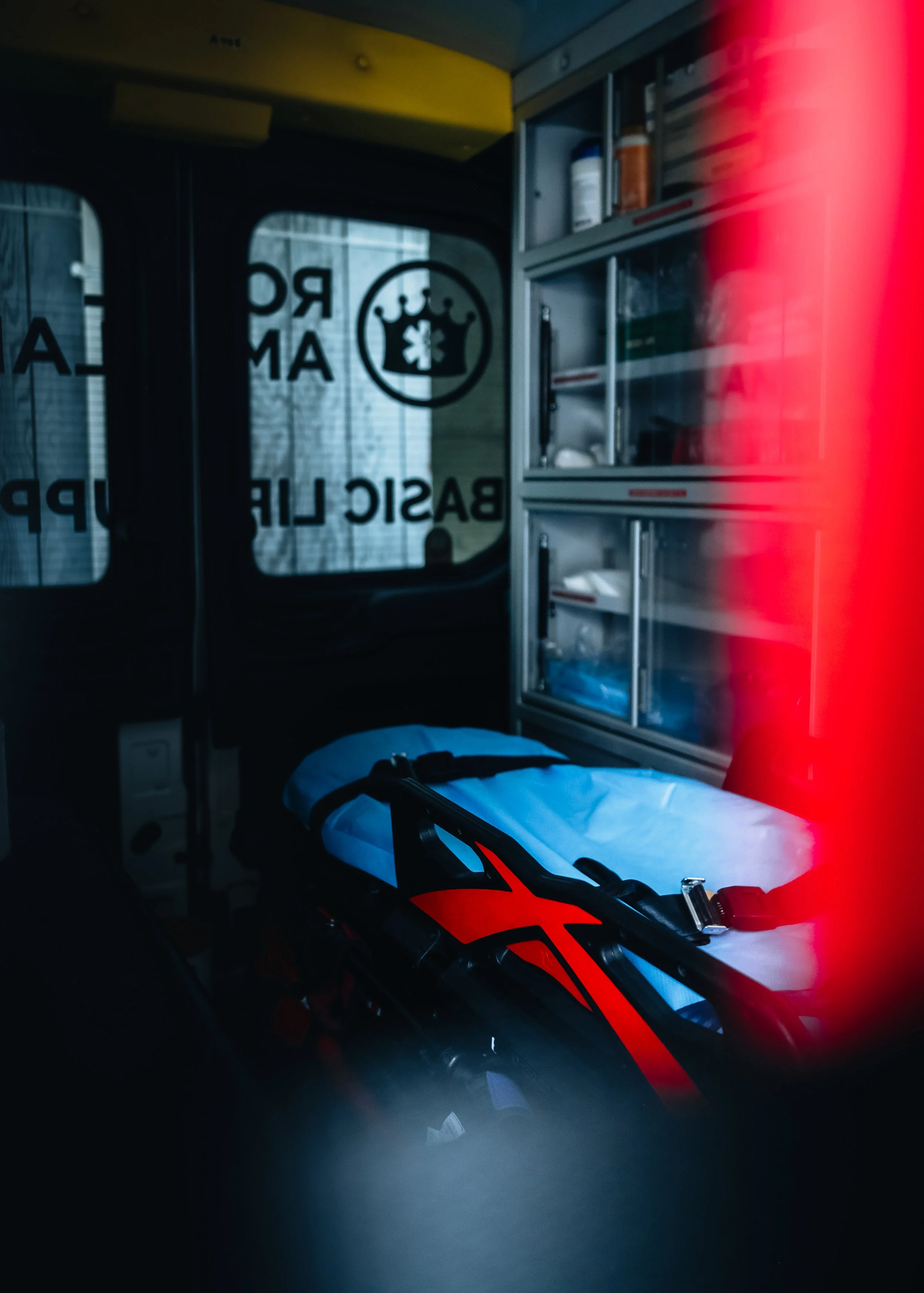 Inside an ambulance, a stretcher with a black and red helmet on top, medical supplies in a cabinet, and a window with a medical emblem and text that reads 'Emergency Medical Response'.
