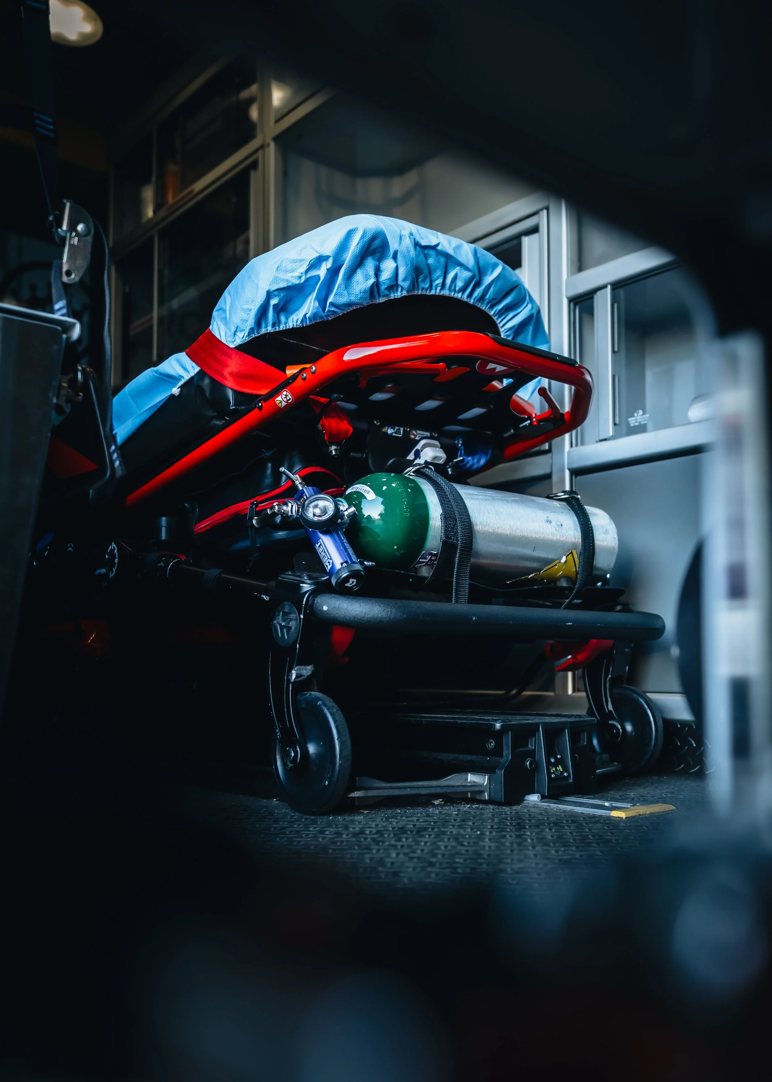 A stretcher with medical supplies in an emergency vehicle or ambulance, including a green oxygen tank, a blue medical bag, and a red item, in a dimly lit environment.