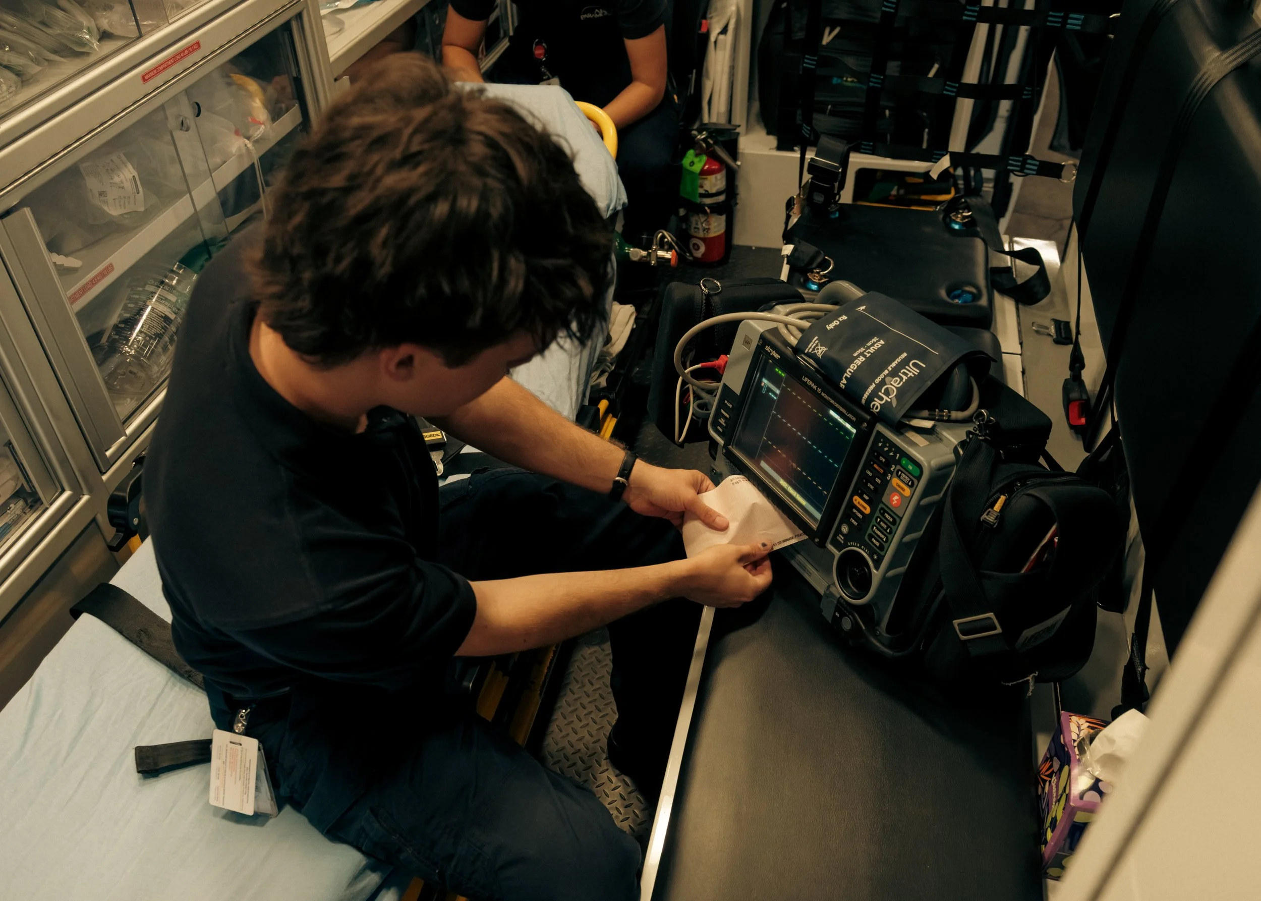 Individual performing medical procedure on patient in ambulance or emergency vehicle using monitoring equipment and supplies.