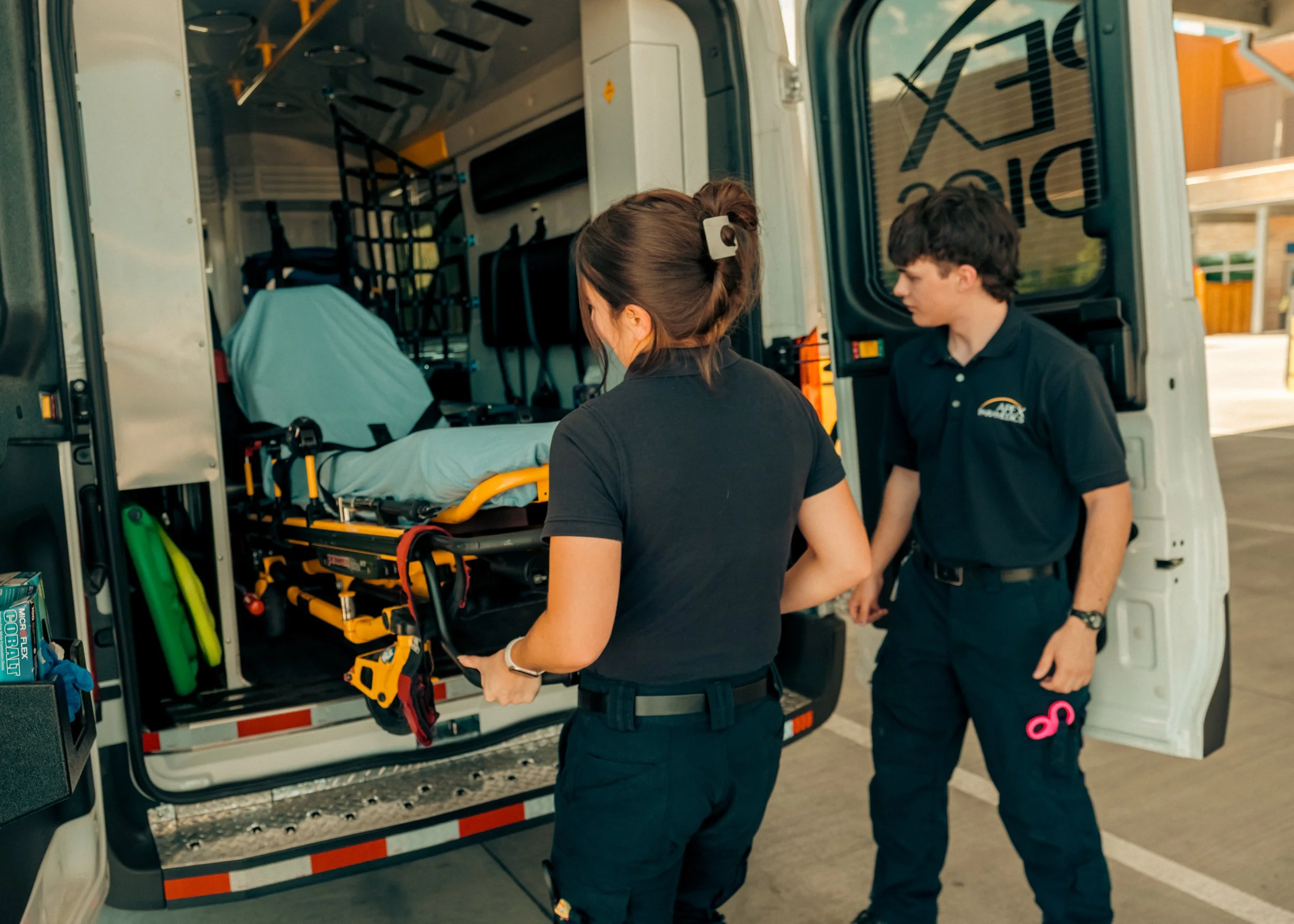 Two emergency medical personnel standing next to an ambulance, preparing medical equipment inside the ambulance.