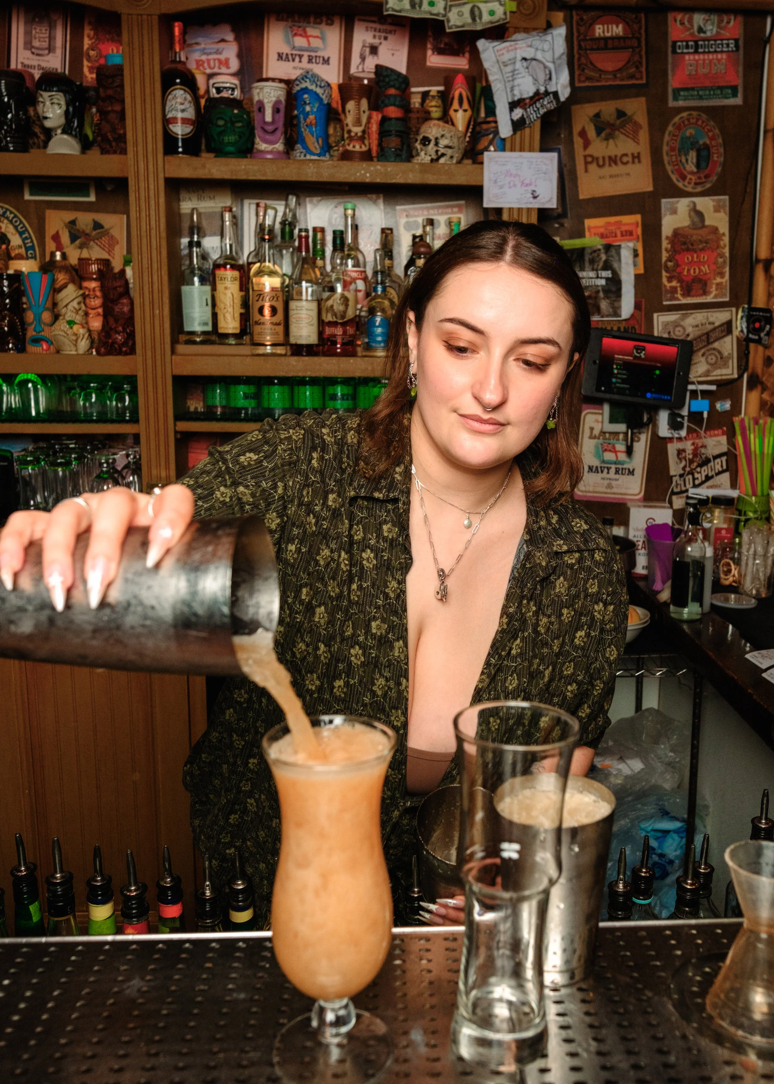 A woman with shoulder-length brown hair pouring a drink into a cocktail glass at a bar surrounded by colorful liquor bottles, posters, and decorations.