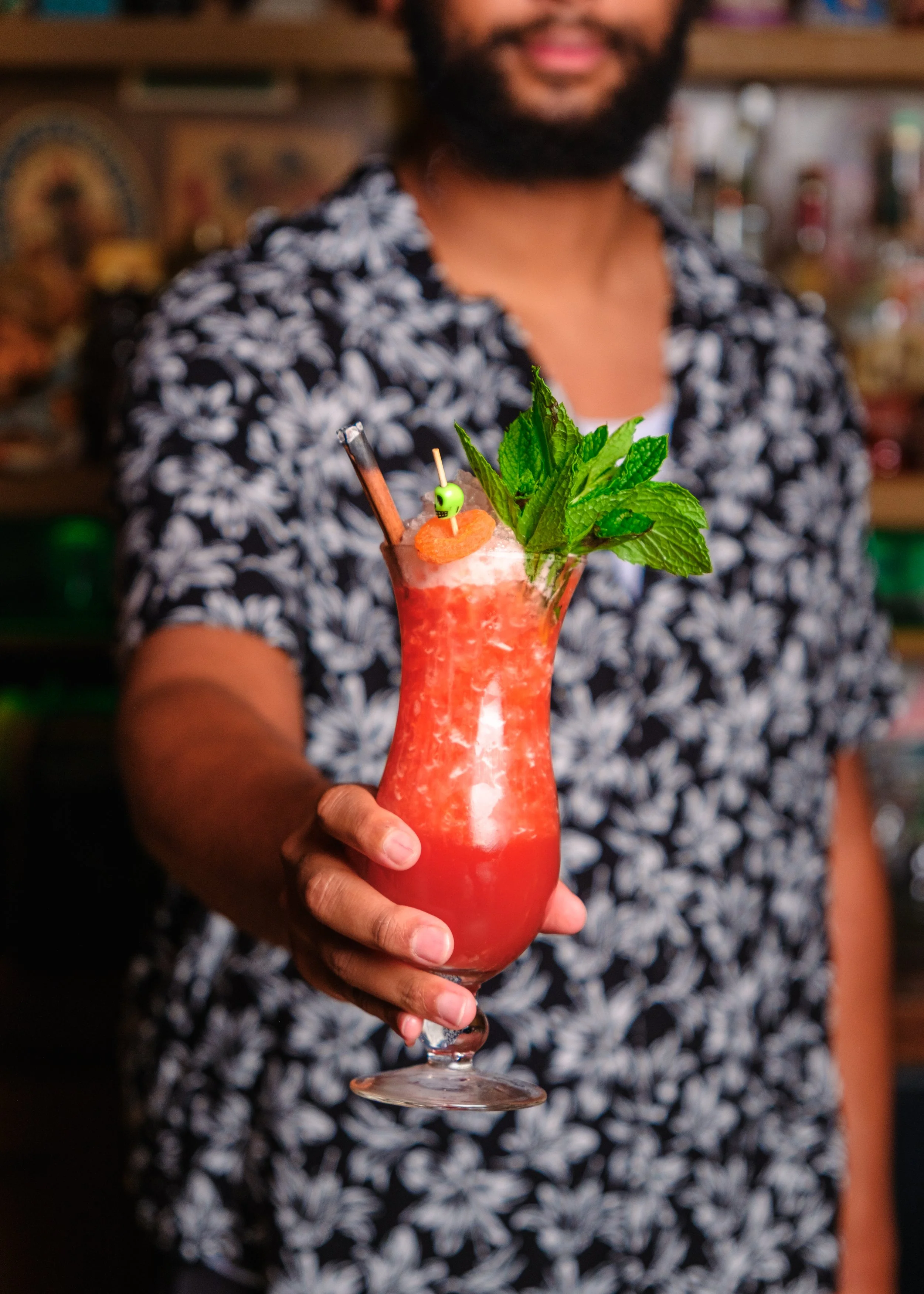 A person holding a pink cocktail garnished with fresh mint leaves, a slice of orange, and a small green skull pick, with a straw.