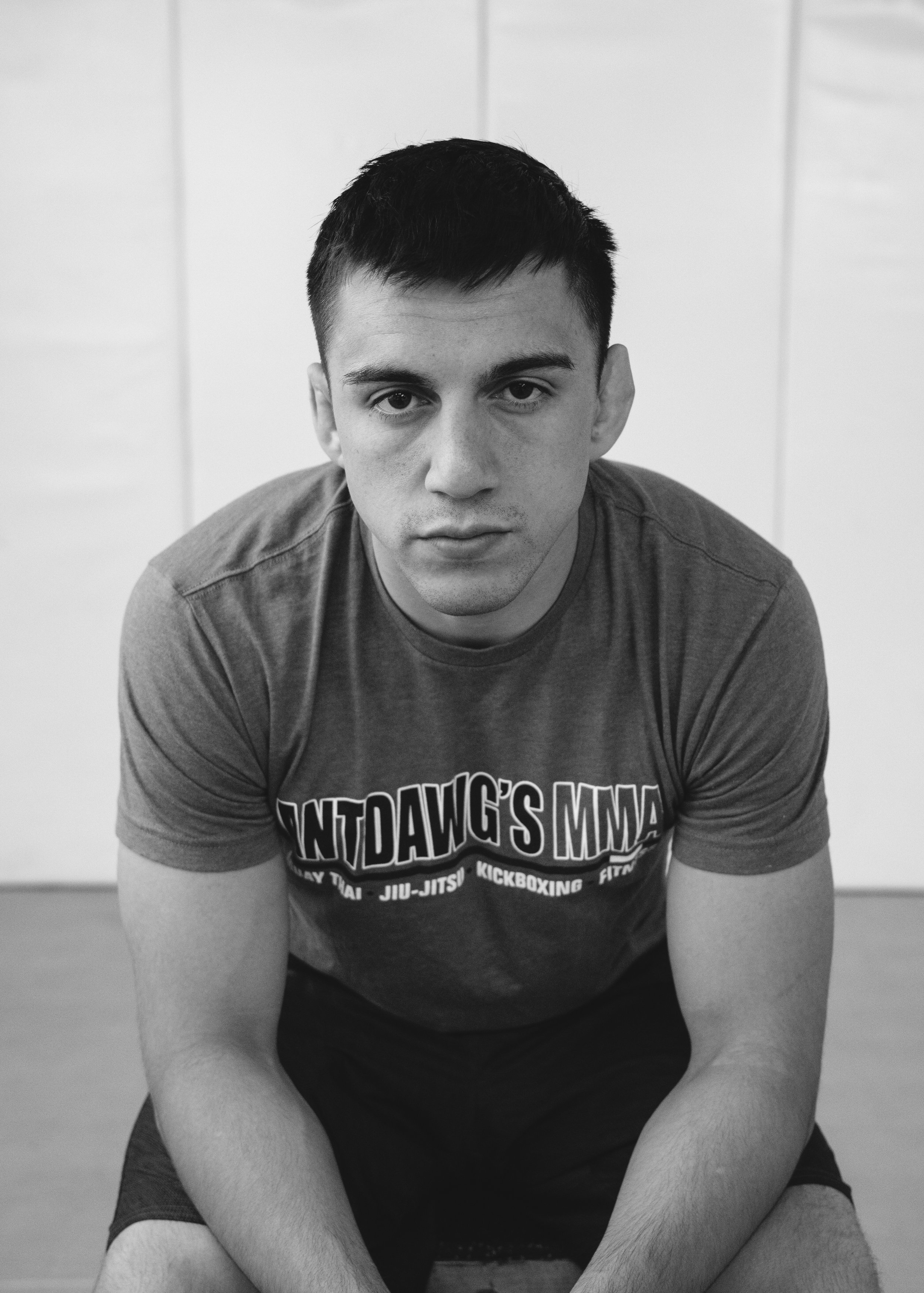 A young man with short dark hair and light skin, sitting and looking directly at the camera, wearing a dark T-shirt with text related to martial arts. Black and white photograph.