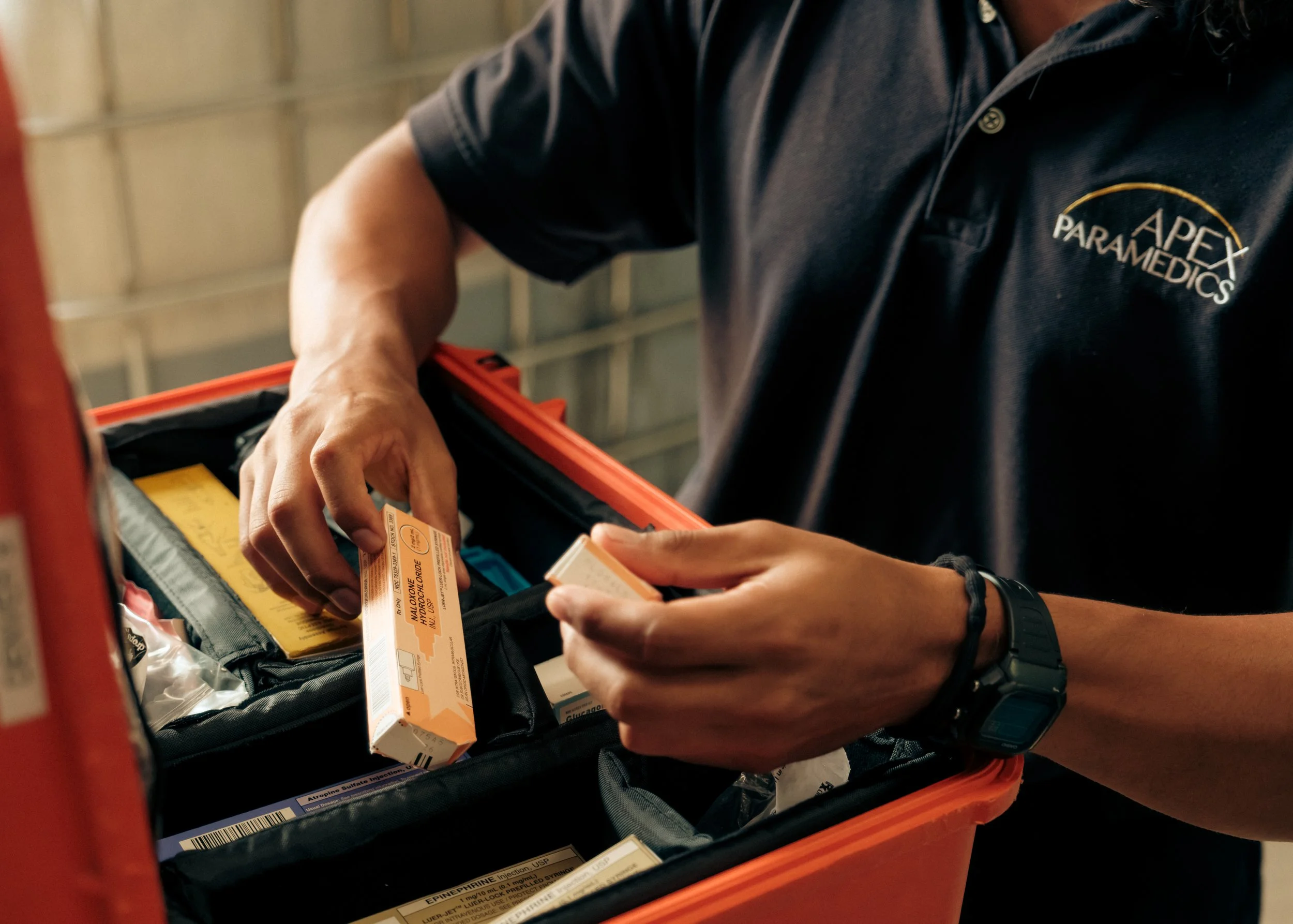 A person wearing a black shirt with a logo that reads 'APEX PARAMEDICS' is reaching into a black bag, holding a box of medication or medical supplies.