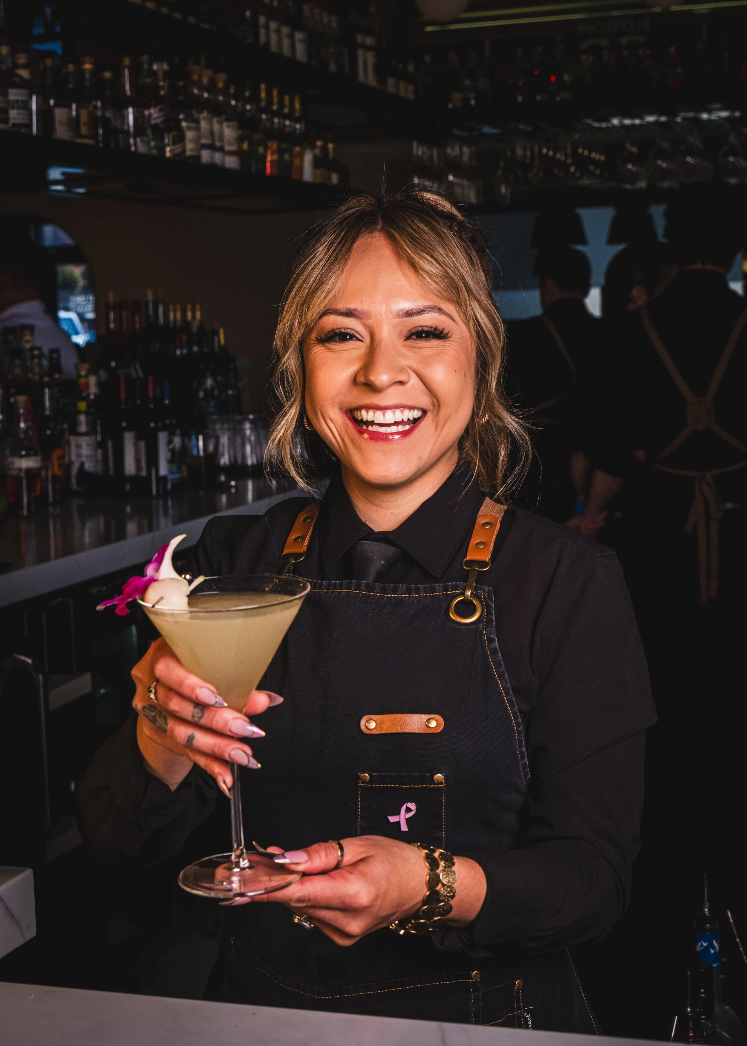 A woman with short blonde hair smiling and holding a cocktail glass in a bar, wearing a black shirt with leather shoulder accents, jewelry, and a pink ribbon sticker