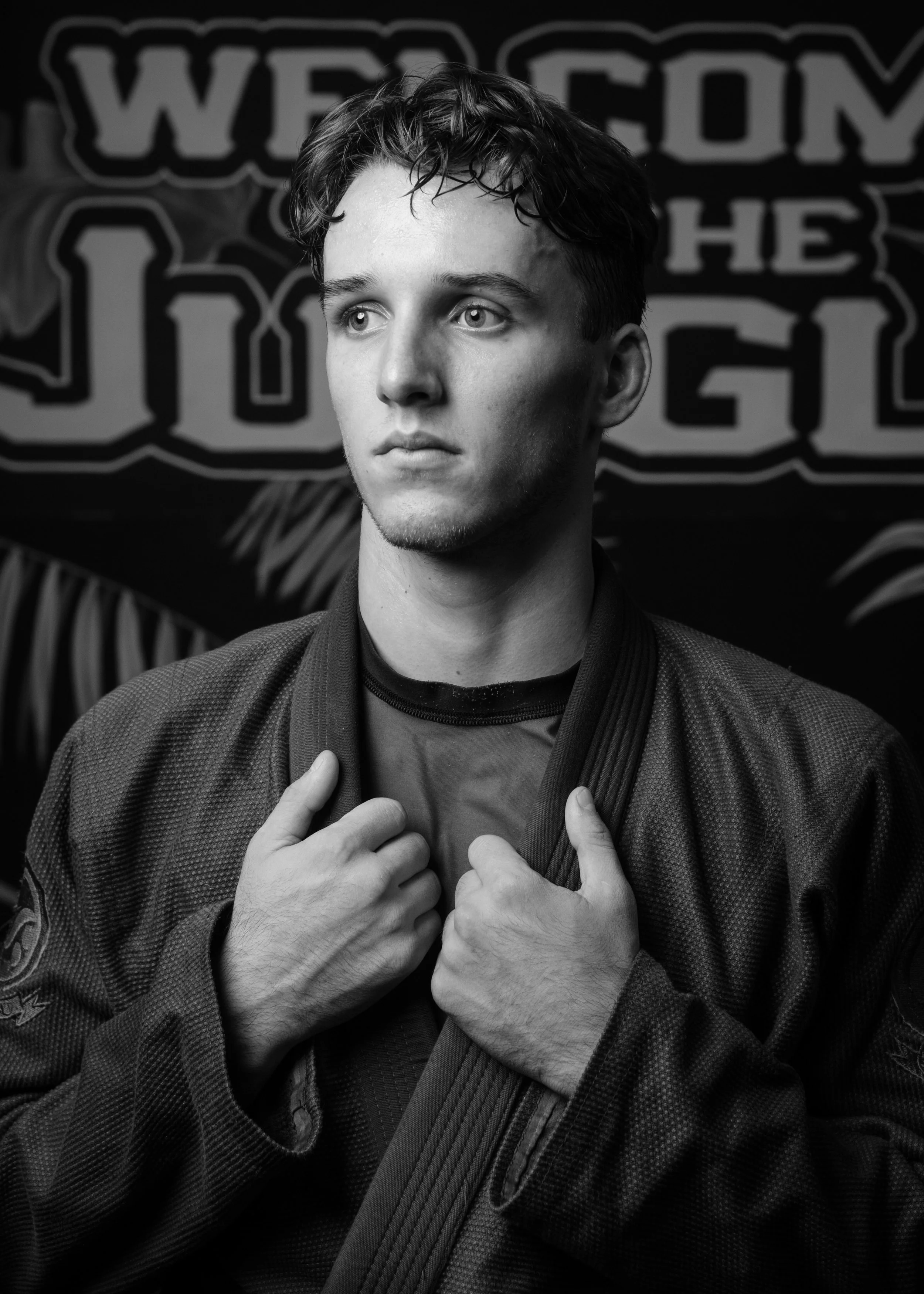 Black and white photo of a young man with short, wet hair wearing a jiu-jitsu gi, holding the collar of his gi with both hands. There is a blurred background with a sign that says "WECOM" and some decorative palm leaves.