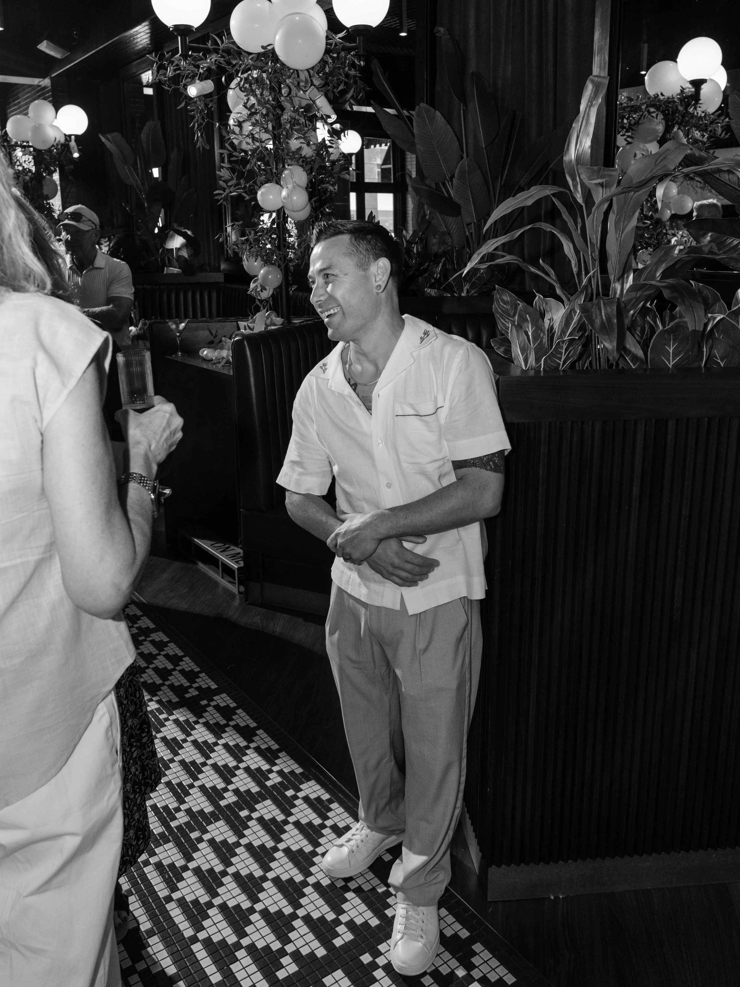 A man smiling while talking to a woman in a restaurant with decorative plants and hanging spherical lights