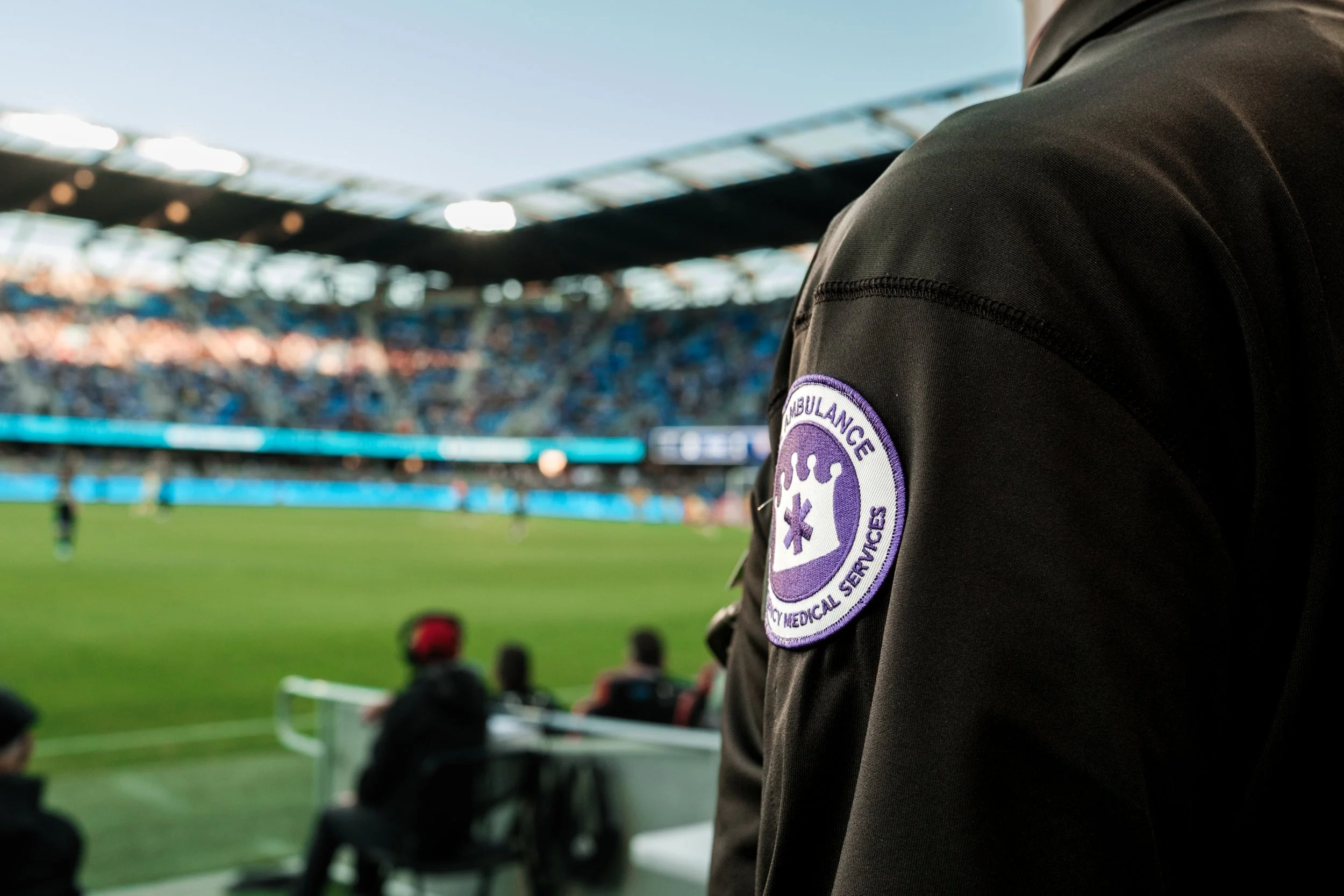 Close-up of a person wearing a black jacket with a patch for emergency medical services, standing near a soccer field with spectators in a stadium in the background.