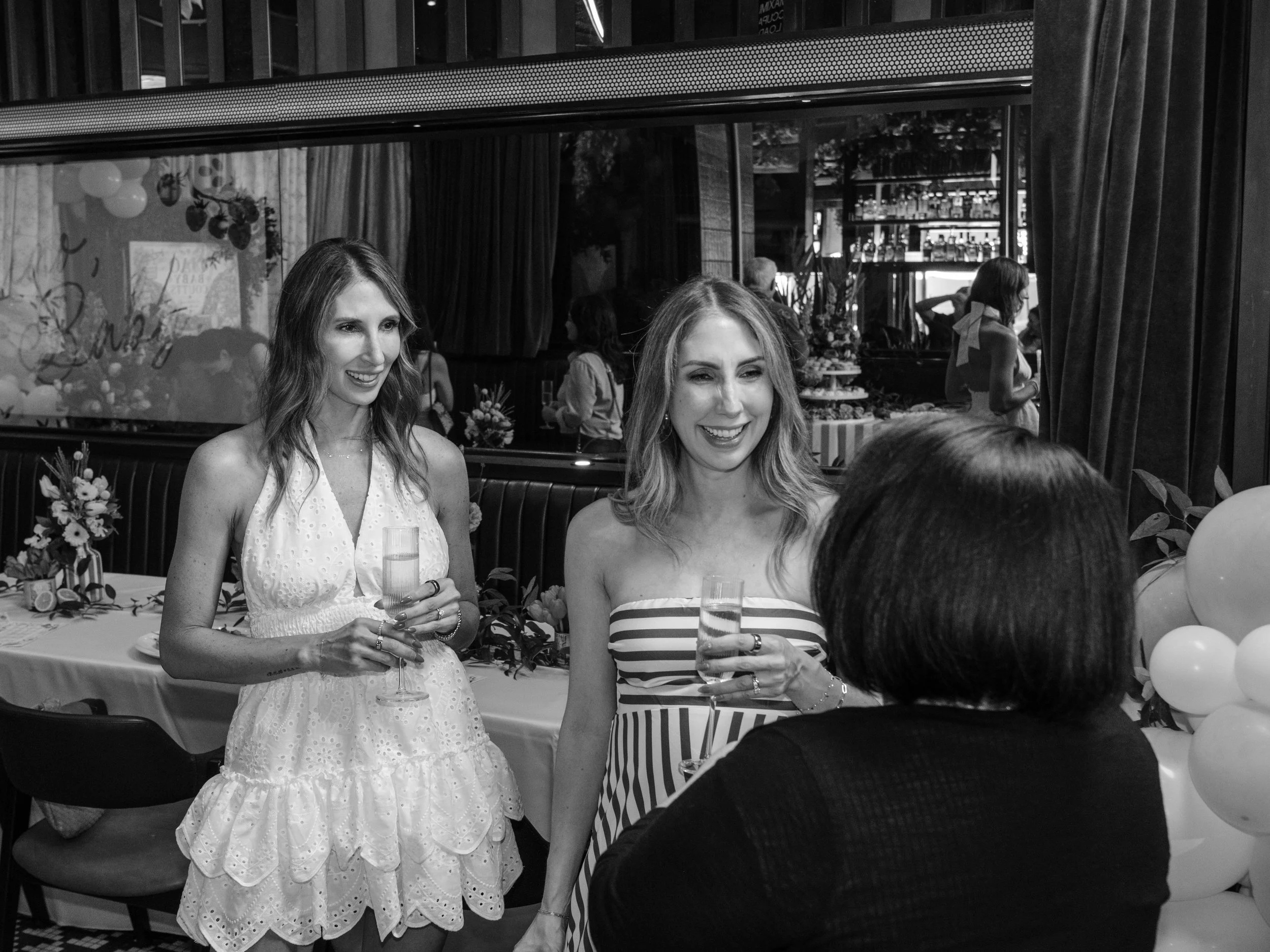 Two smiling women holding glasses of champagne at a celebration or party. They are standing in front of a decorated table with balloons and flowers, with other people in the background.