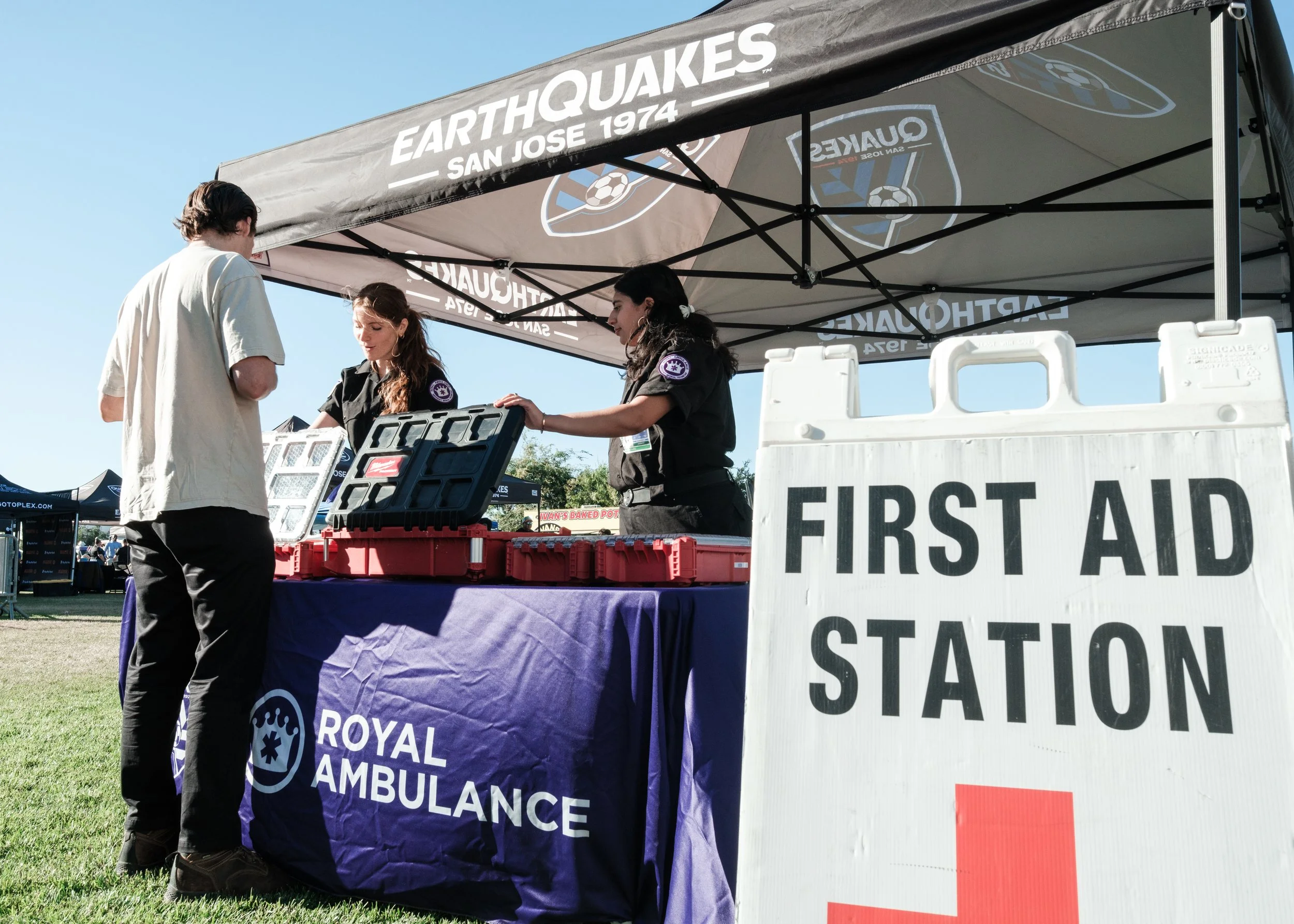 Outdoor scene featuring a person with dark hair and light-colored clothing talking to two emergency medical personnel under a black canopy tent that reads "Earthquakes San Jose 1974". The scene includes a table covered with a purple cloth displaying 