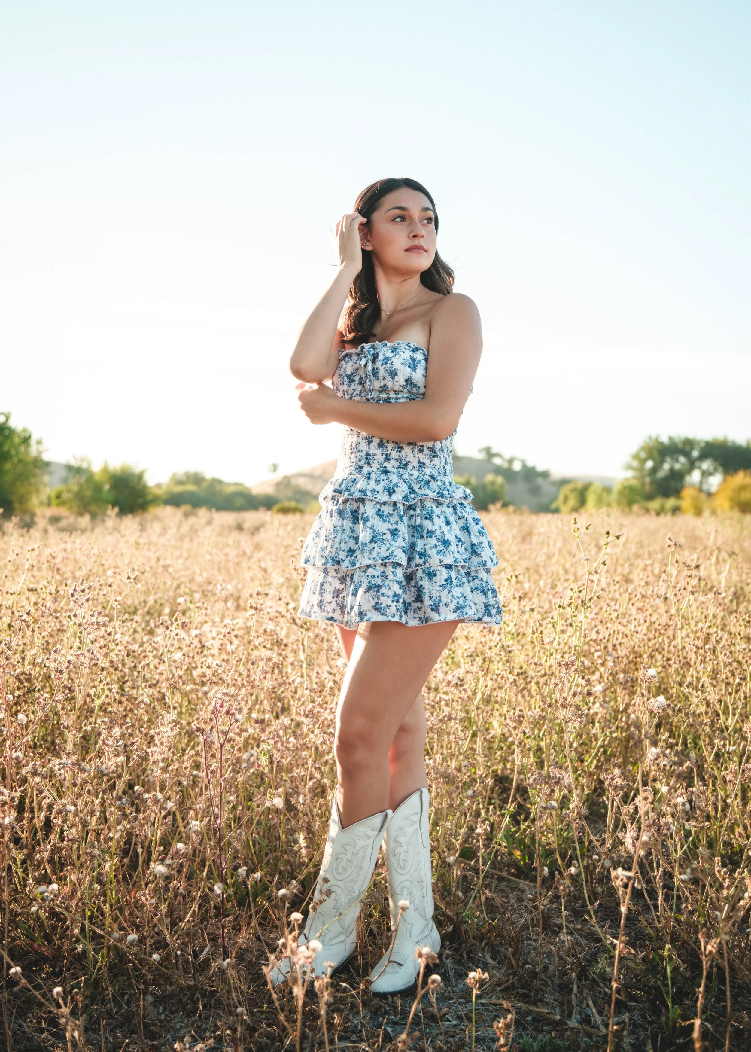 A young woman standing in a field of pinkish-brown wildflowers under a clear sky, wearing a blue floral dress, white cowboy boots, and touching her hair with one hand.