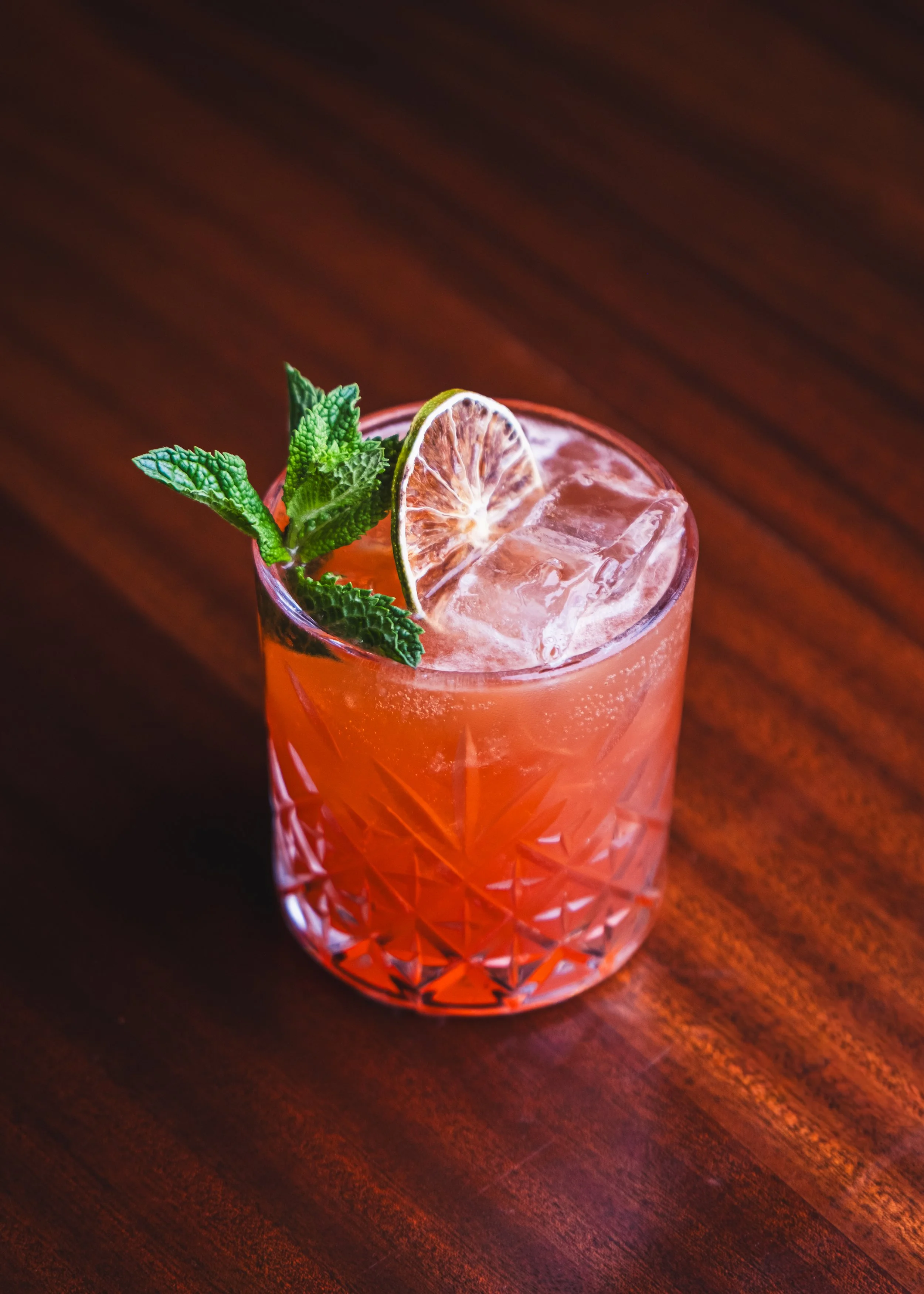 A glass of pinkish cocktail garnished with a slice of dried lime and a sprig of fresh mint, on a dark wooden surface.