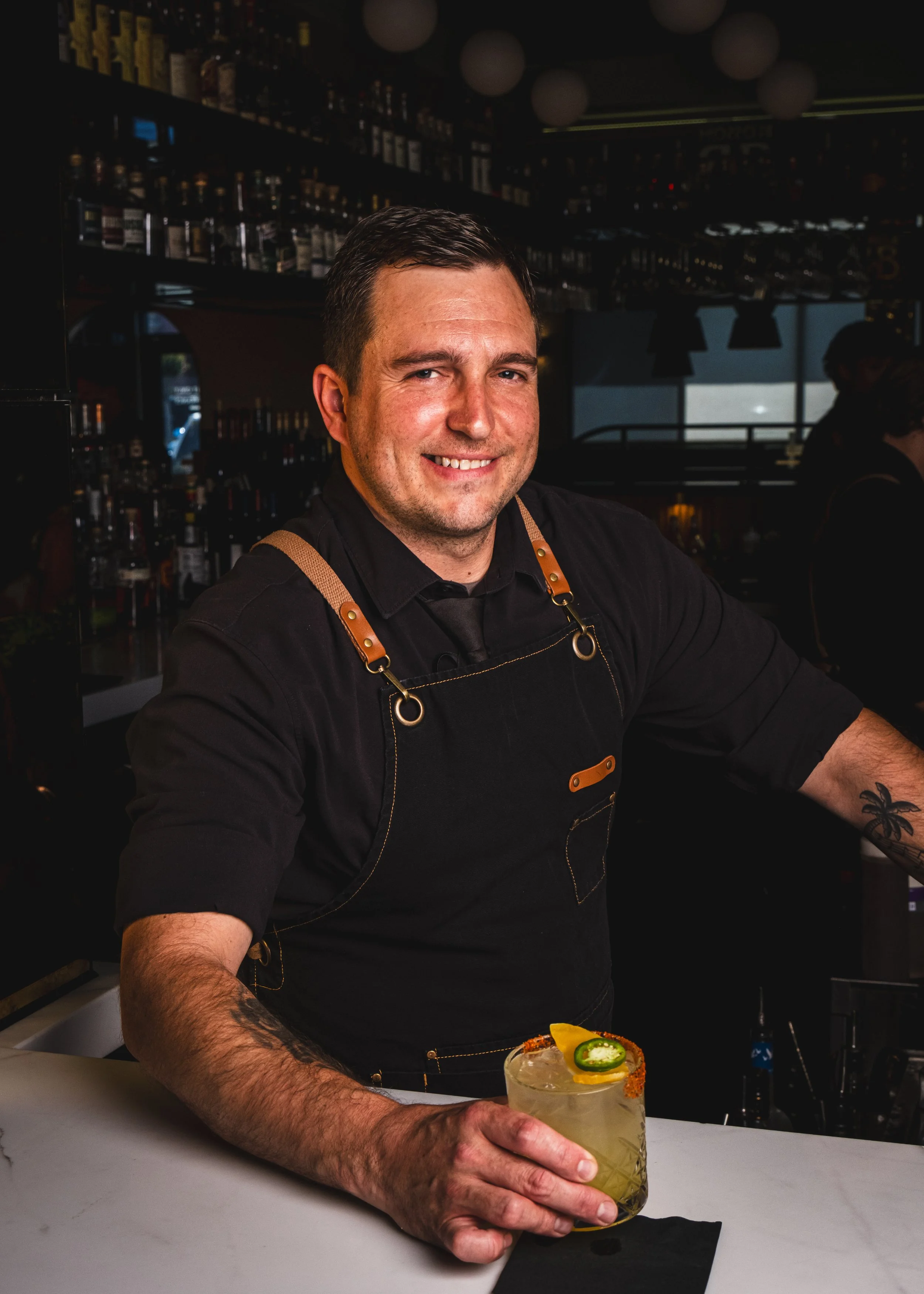 A bartender with short dark hair, wearing a black shirt and brown apron, is smiling and holding a cocktail glass with a jalapeño slice, lemon wedge, and salted rim on a white bar counter. The bar has bottles and glasses in the background under dim li