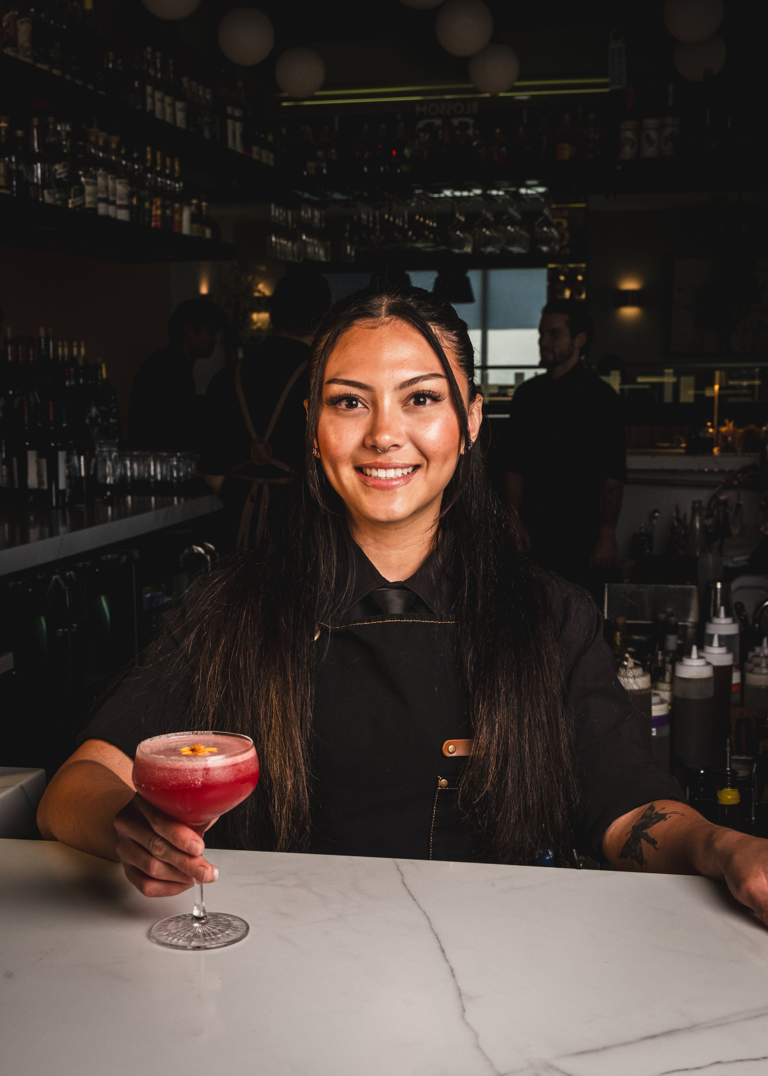 A woman with long dark hair, smiling, holding a pink cocktail with a flower garnish at a bar. She wears a black top and has a tattoo on her arm. The bar has a marble countertop and a dark, busy background with shelves of bottles and bartenders.