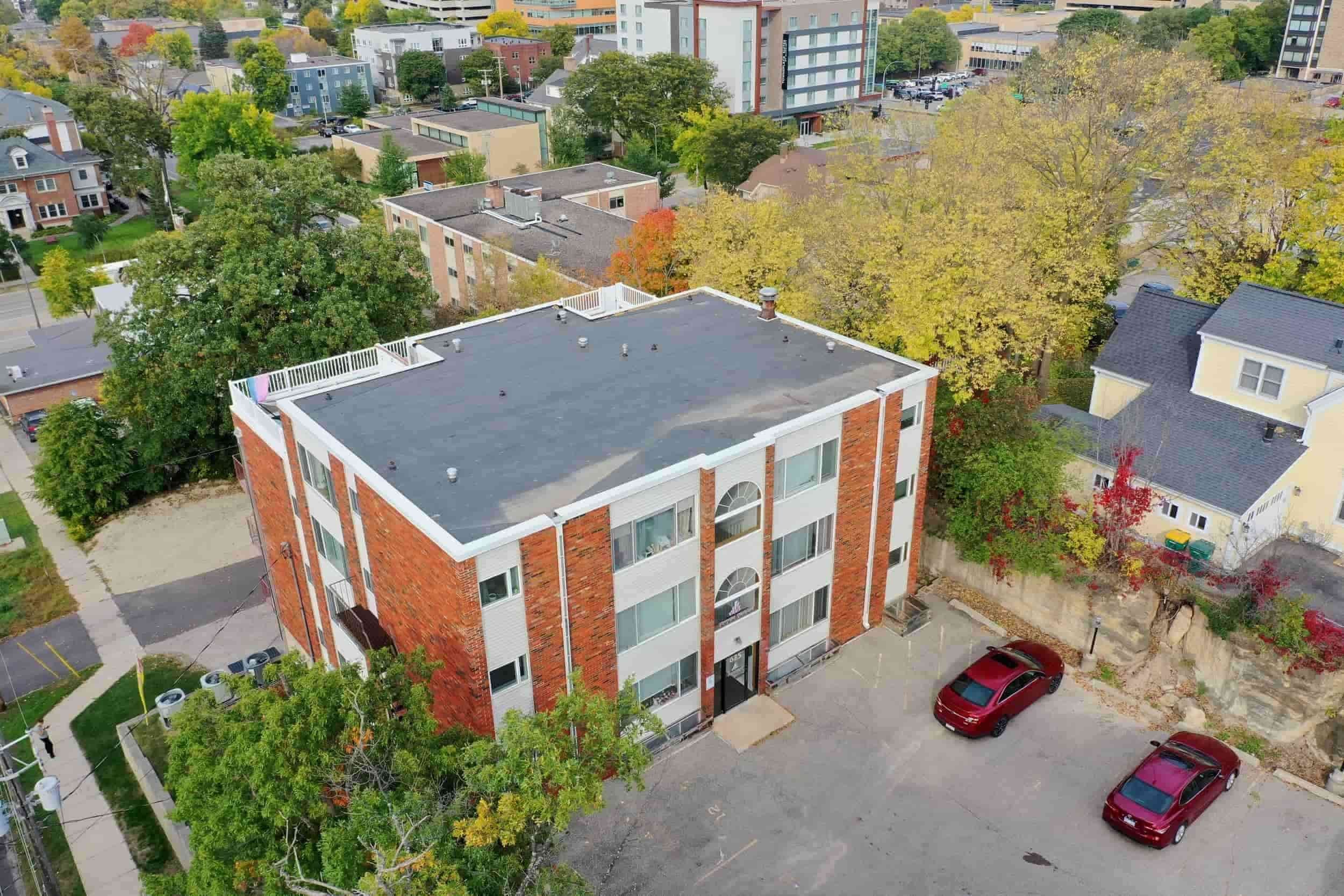 Aerial view of apartments in Rochester: three-story red brick building, parking lot with a red car, trees, and cityscape.