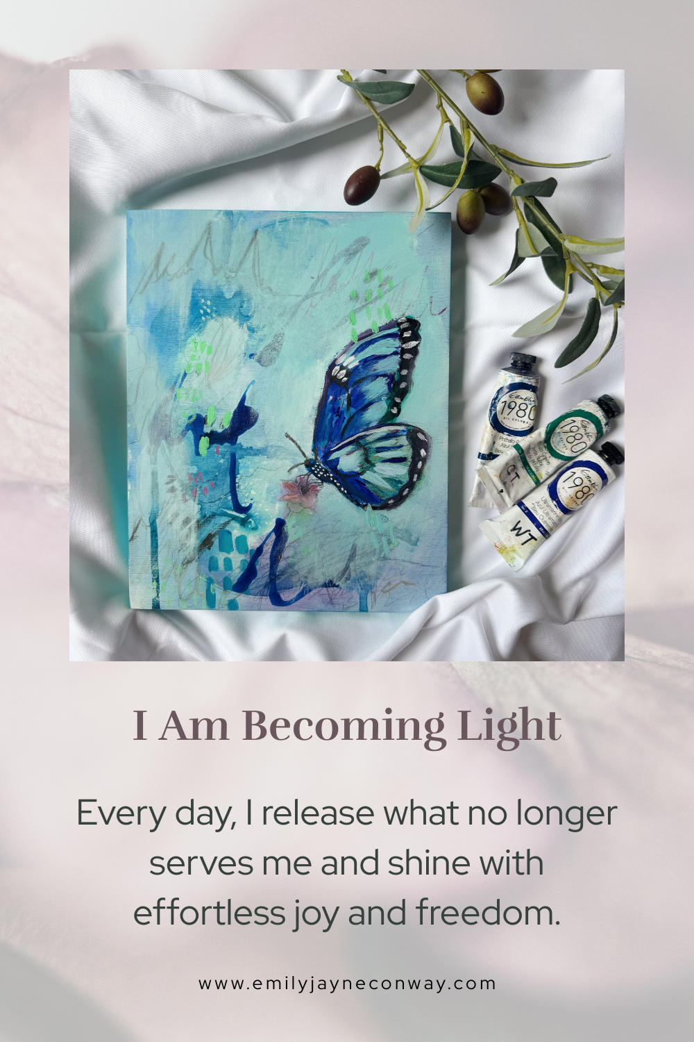 An abstract watercolor painting of a blue butterfly on a canvas, surrounded by three tubes of watercolor paint and some olive leaves on a silky white fabric.
