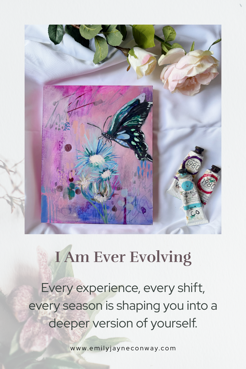 A painting of a butterfly and flowers on a pink background, surrounded by white roses and three tubes of watercolor paint on a white fabric surface.