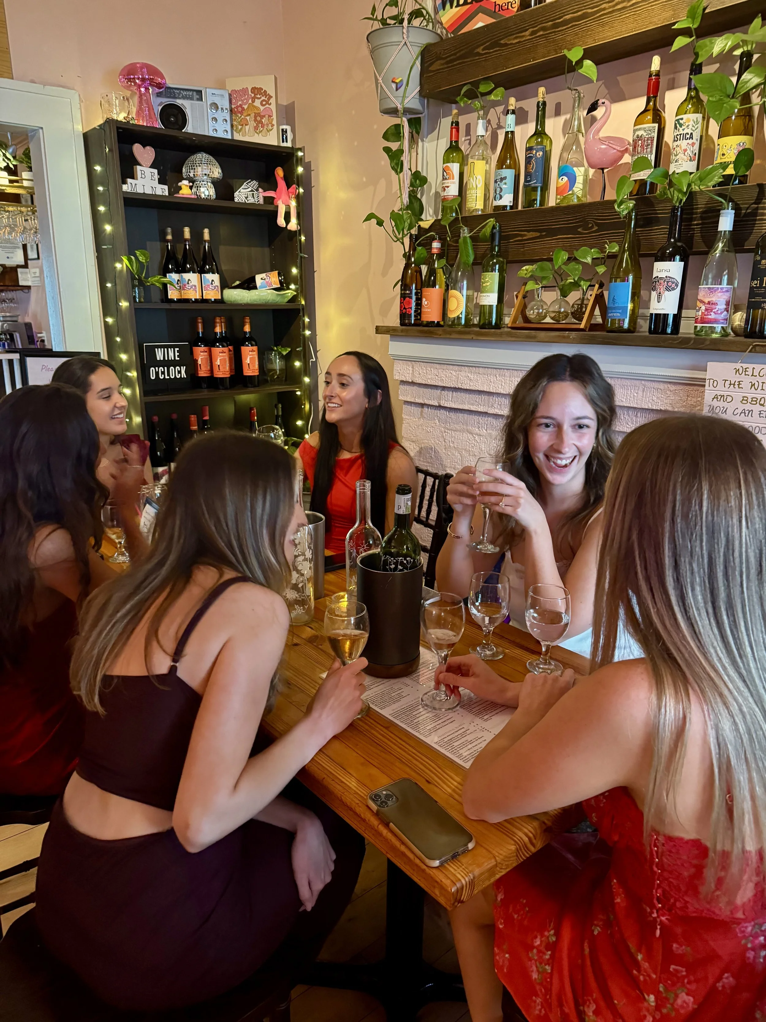 Group of women sitting around a wooden table in a cozy restaurant, smiling and enjoying drinks. The background features shelves with colorful bottles of wine and decorative items, including a pink flamingo statue and potted plants.