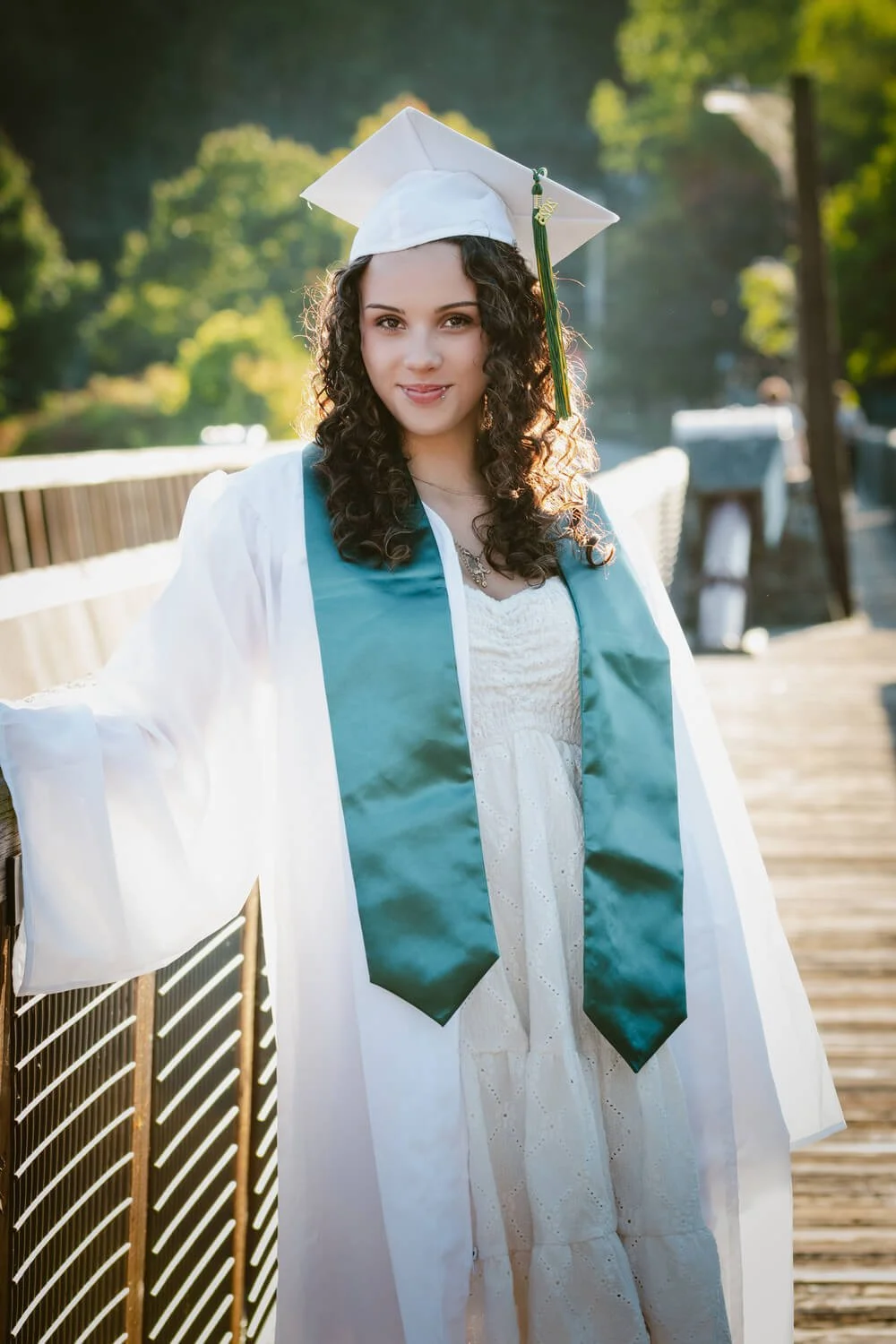 Young woman in a white graduation gown and cap with a teal sash, standing outdoors on a wooden walkway, smiling, with trees and sunlight in the background.