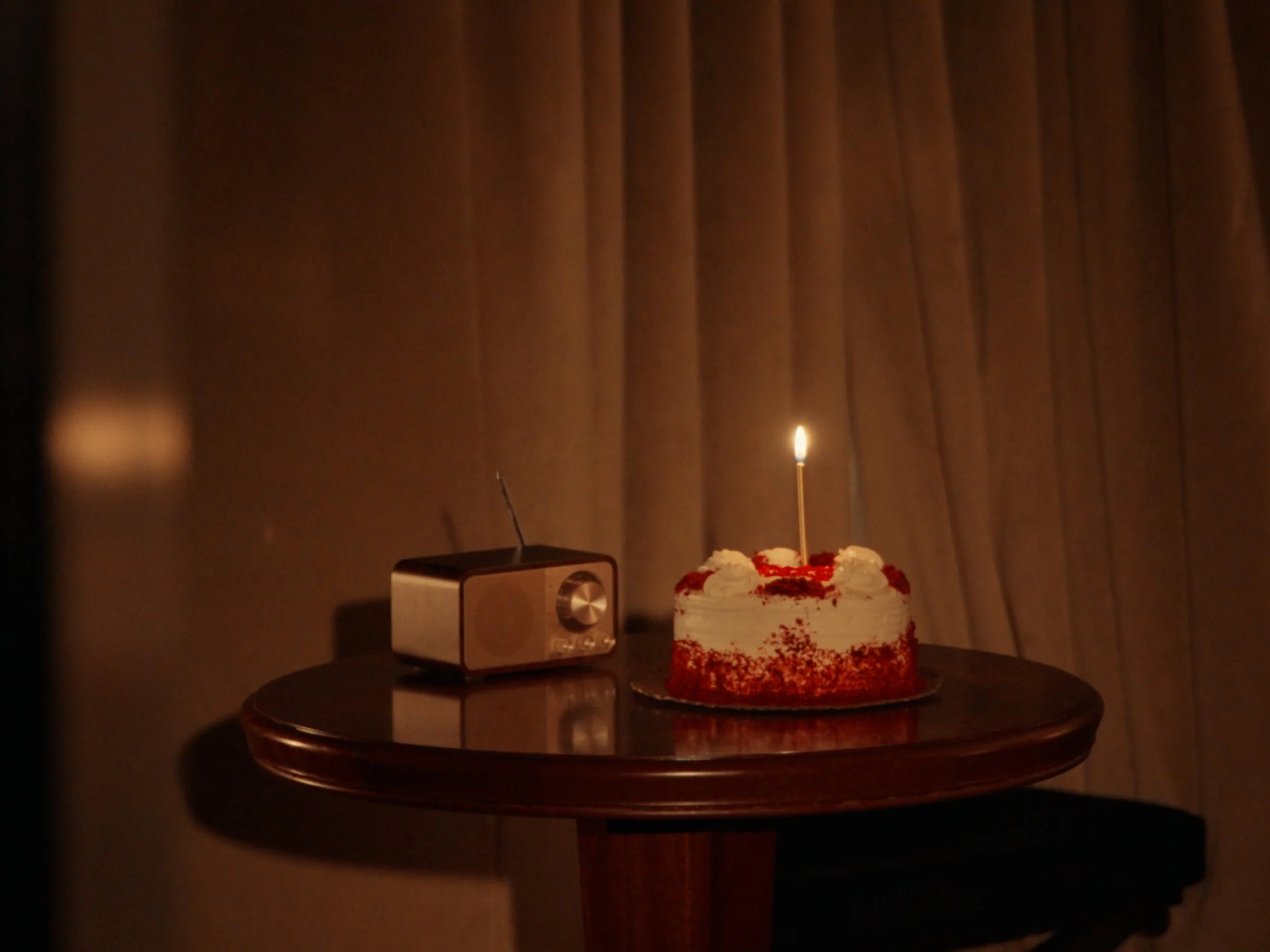 A birthday cake with white frosting and red sprinkles, topped with a single lit candle, on a wooden table beside a black and silver speaker.
