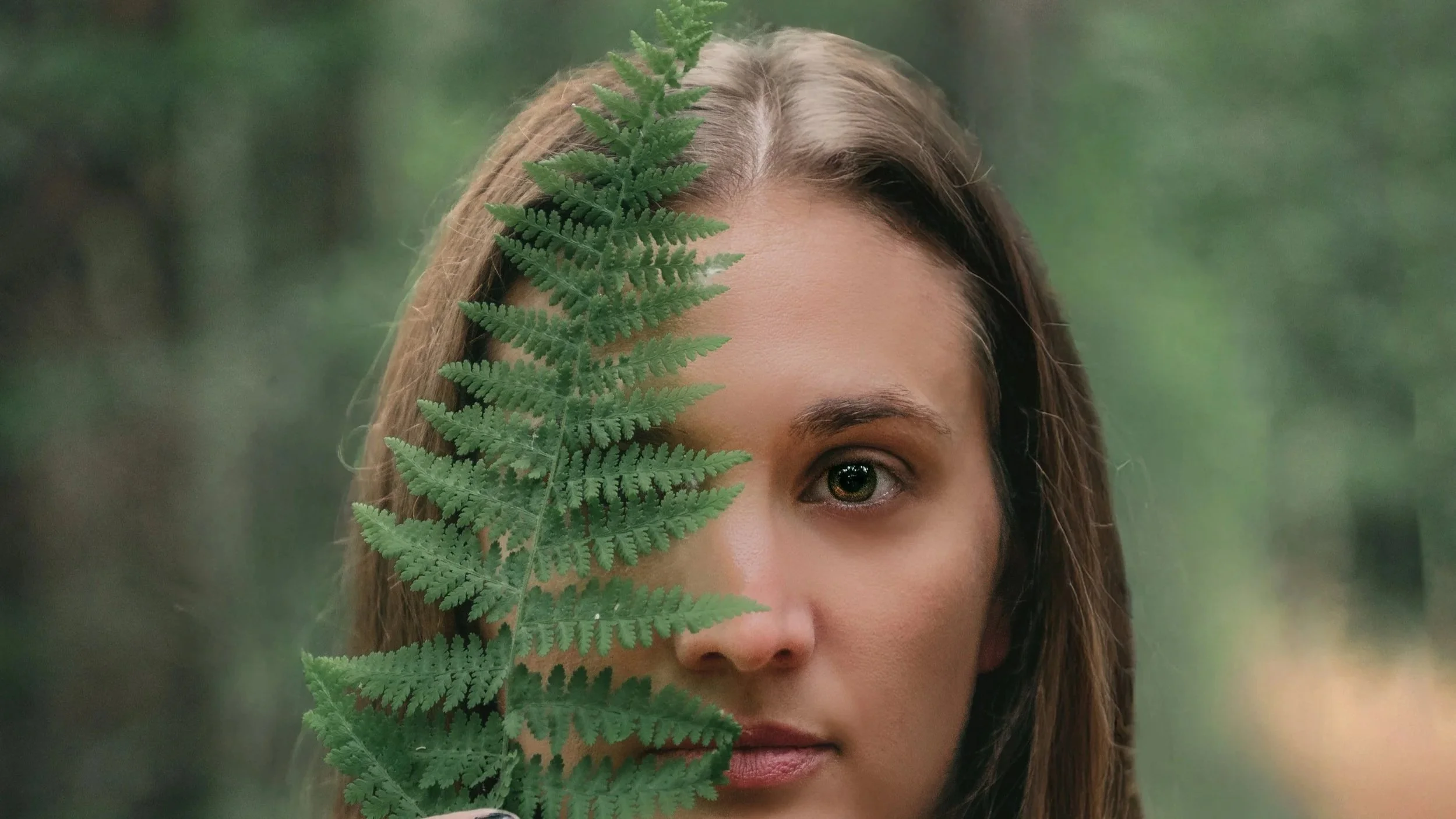 A woman with light skin and brown hair holds a green fern leaf in front of her face, covering her forehead and part of her right eye, with a blurred green outdoor background.