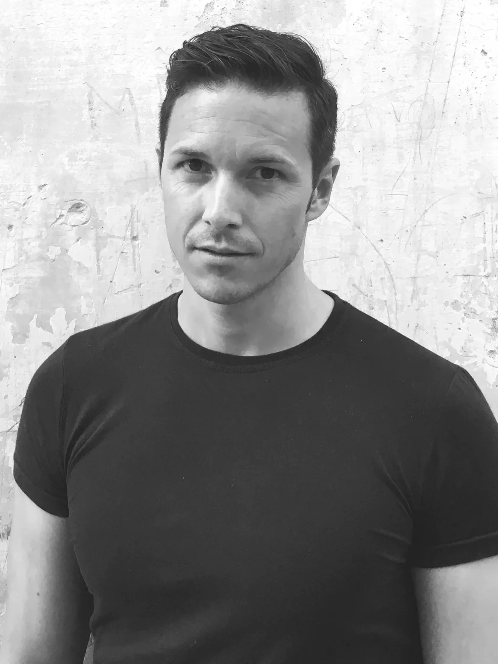 Schwarz-weiß-Foto eines Mannes mit dunklem, kurzer, gewelltem Haar, blickt leicht zu Seite, trägt ein schwarzes T-Shirt, im Hintergrund eine weiße, verputzte Wand.