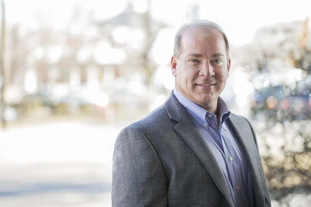 A professional man wearing a gray blazer and blue dress shirt, smiling outdoors with trees and buildings in the background.