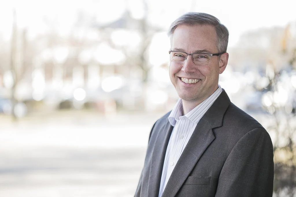 A smiling man wearing glasses and a suit standing outdoors with a blurred background of trees and buildings.