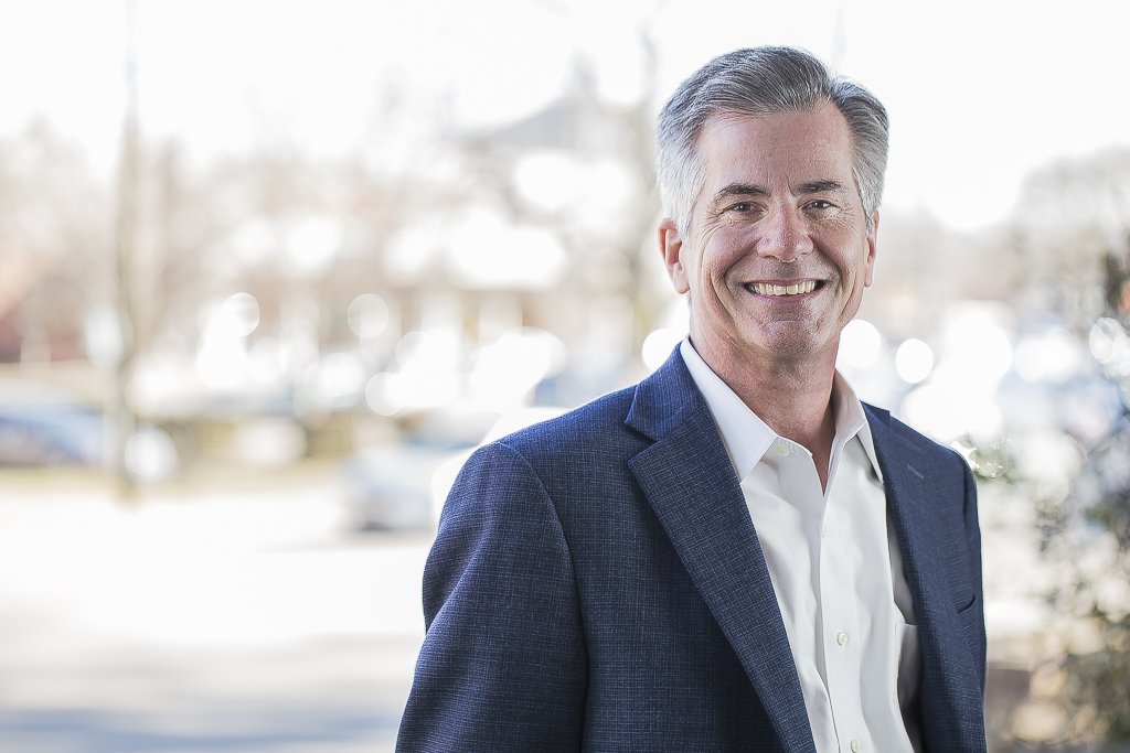 A middle-aged man with gray hair, wearing a dark blue suit and white dress shirt, smiling outdoors with a blurred background of trees and parked cars.