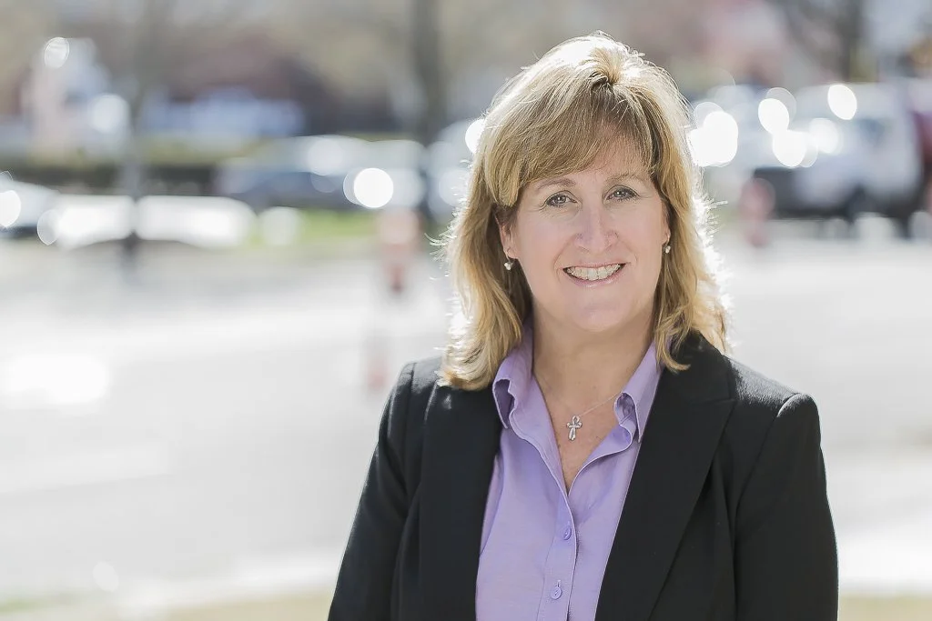 A smiling woman with blonde hair wearing a purple blouse and black blazer stands outdoors with a blurred background of parked cars and trees.