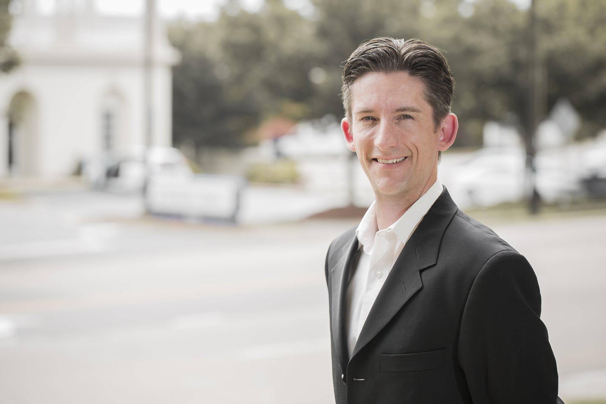 A smiling man in a black suit and white shirt standing outdoors on a city street with blurred background trees and buildings.