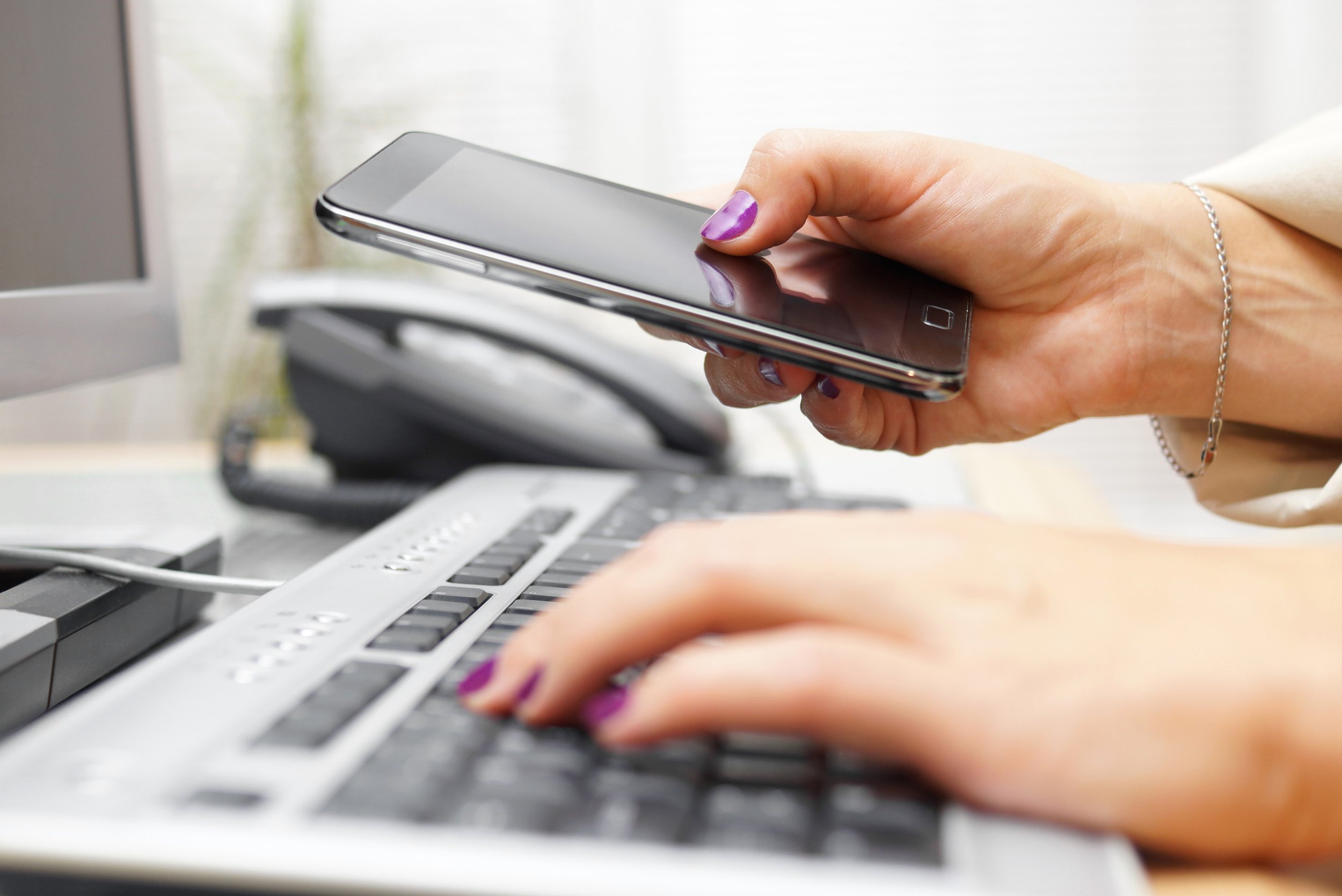 Person using a smartphone while working on a laptop, with a landline phone and a computer monitor nearby.