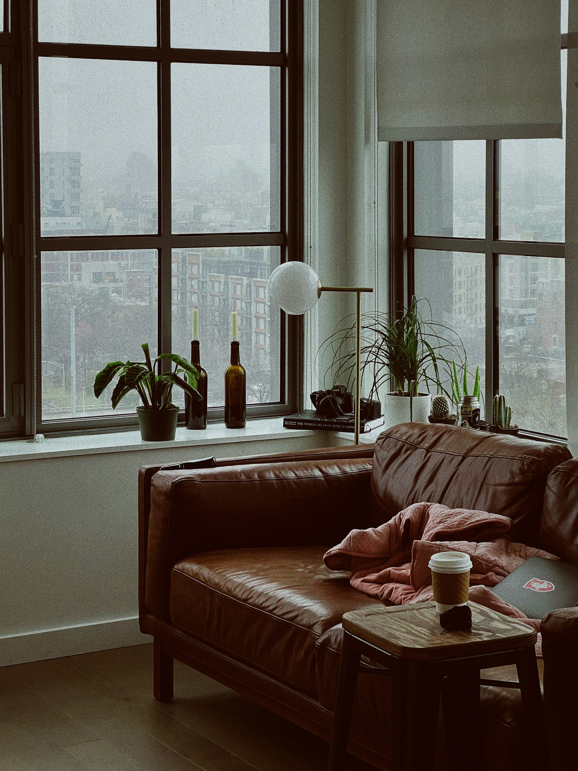 Modern masculine living room with a large window showing a cityscape, a brown leather sofa with a pink blanket and a notebook, a small wooden side table with a coffee cup, potted plants, bottles, a book, and a laptop.
