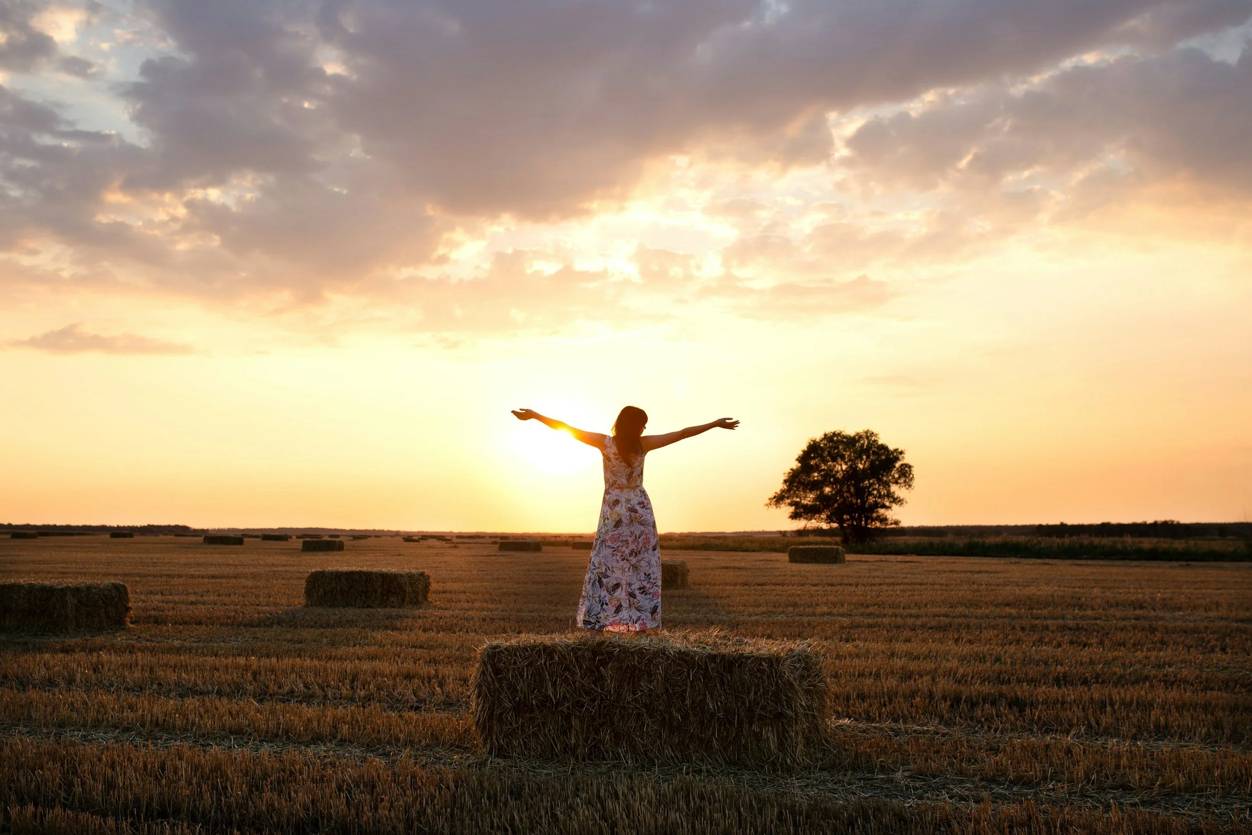 Woman standing on a hay bale with arms outstretched, facing the sun, symbolizing freedom, healing, and emotional renewal.