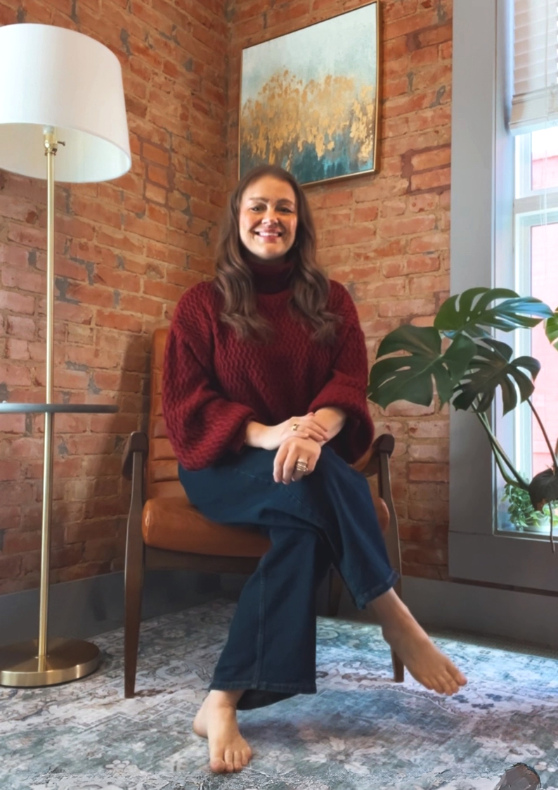 Elizabeth Boyer, LMFT sitting in a brown chair, smiling, with brick walls, a window with blinds, a potted plant, and a framed painting behind her.