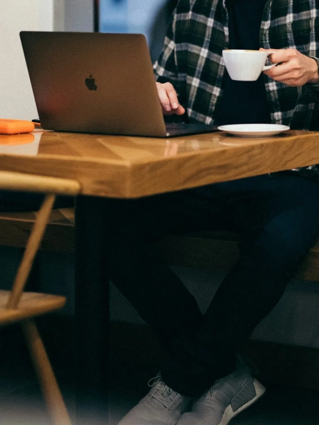 Man sitting at a wooden table using a MacBook laptop, holding a white cup with a saucer nearby, wearing a black shirt and plaid jacket, with only the lower half and hands visible, filling out a requst for a phone consultation for therapy.