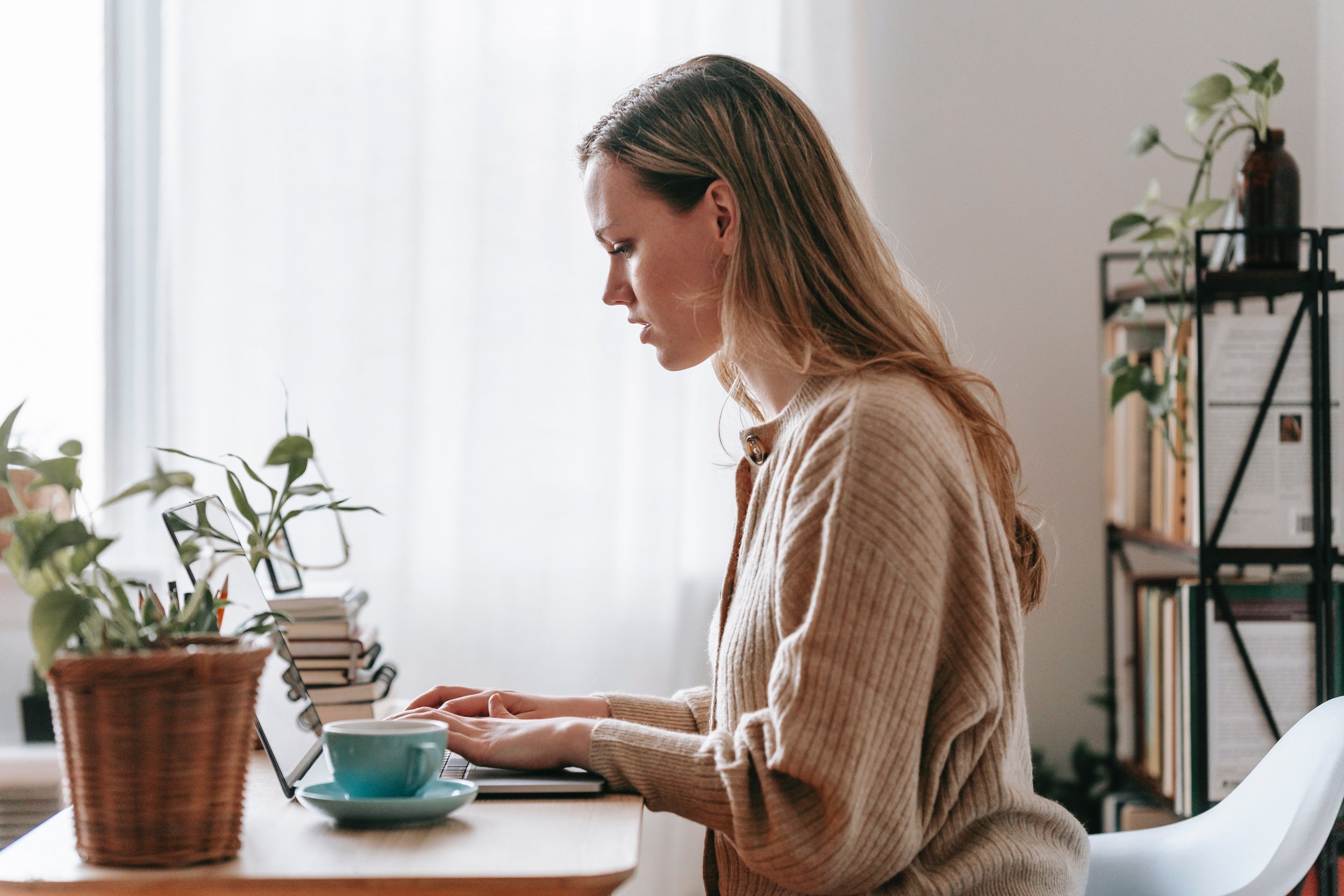 A woman works on a computer with a blue coffee cup on the table next to her.