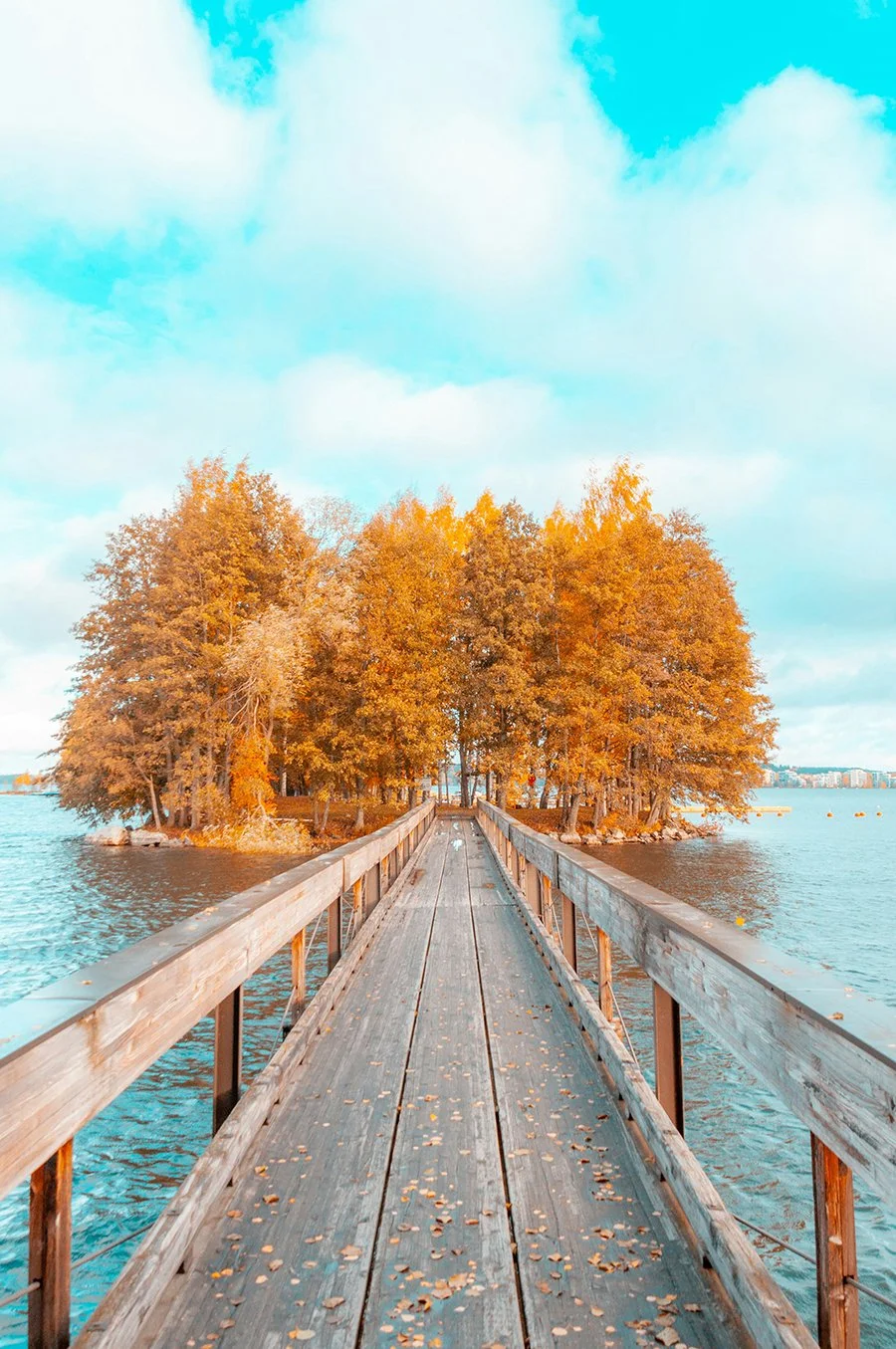 View of a wooden bridge leading to a small island with autumn-colored trees, surrounded by a body of water under a blue sky.