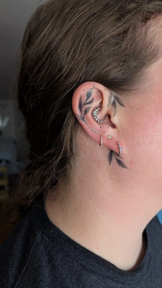 A healed tattoo on a woman's ear. It has leaves in a stipple grey ink, and the tattoo is cutely woven through her earrings.