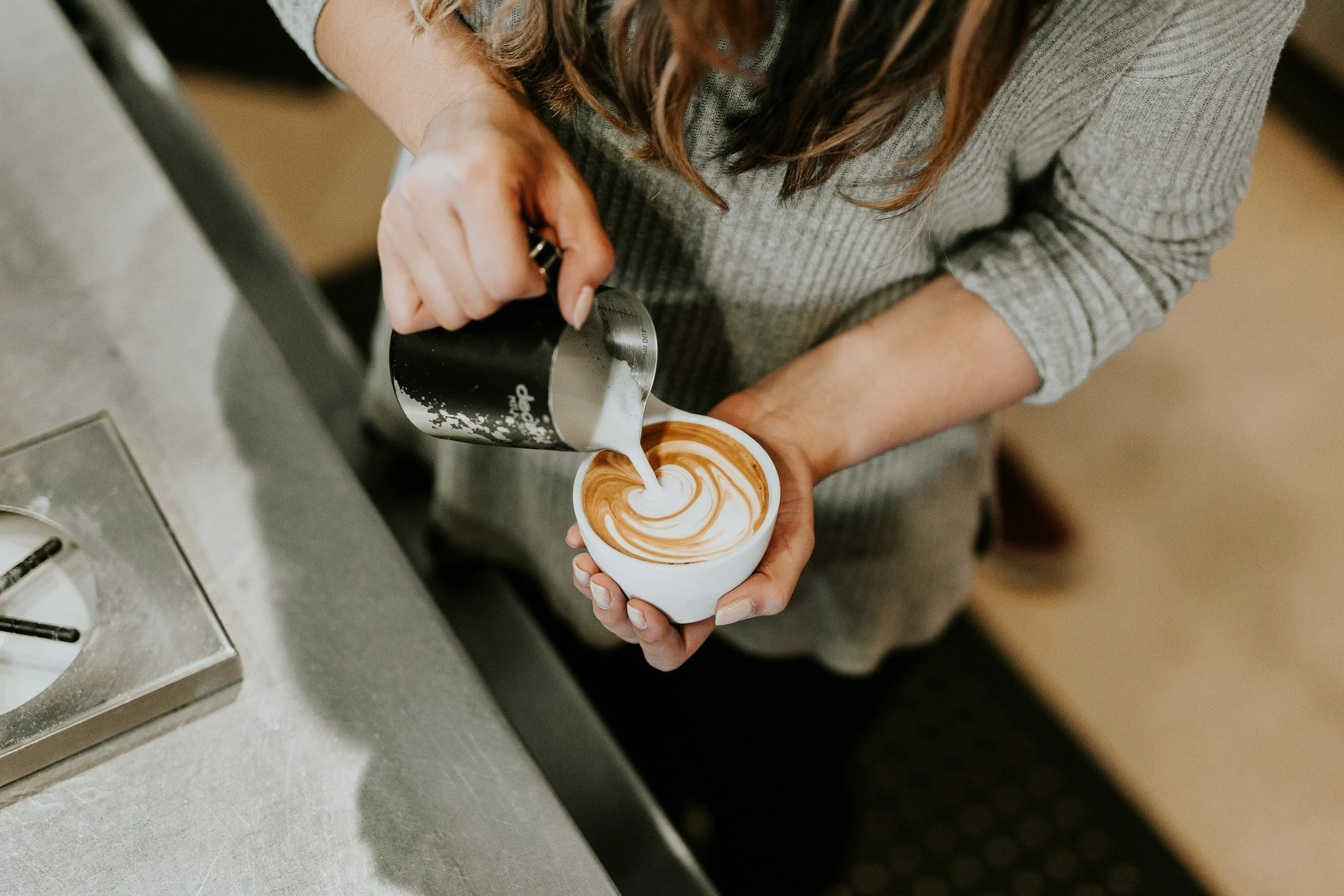 A woman in a gray sweater pouring steamed milk into a cup of espresso to make a latte, creating latte art.