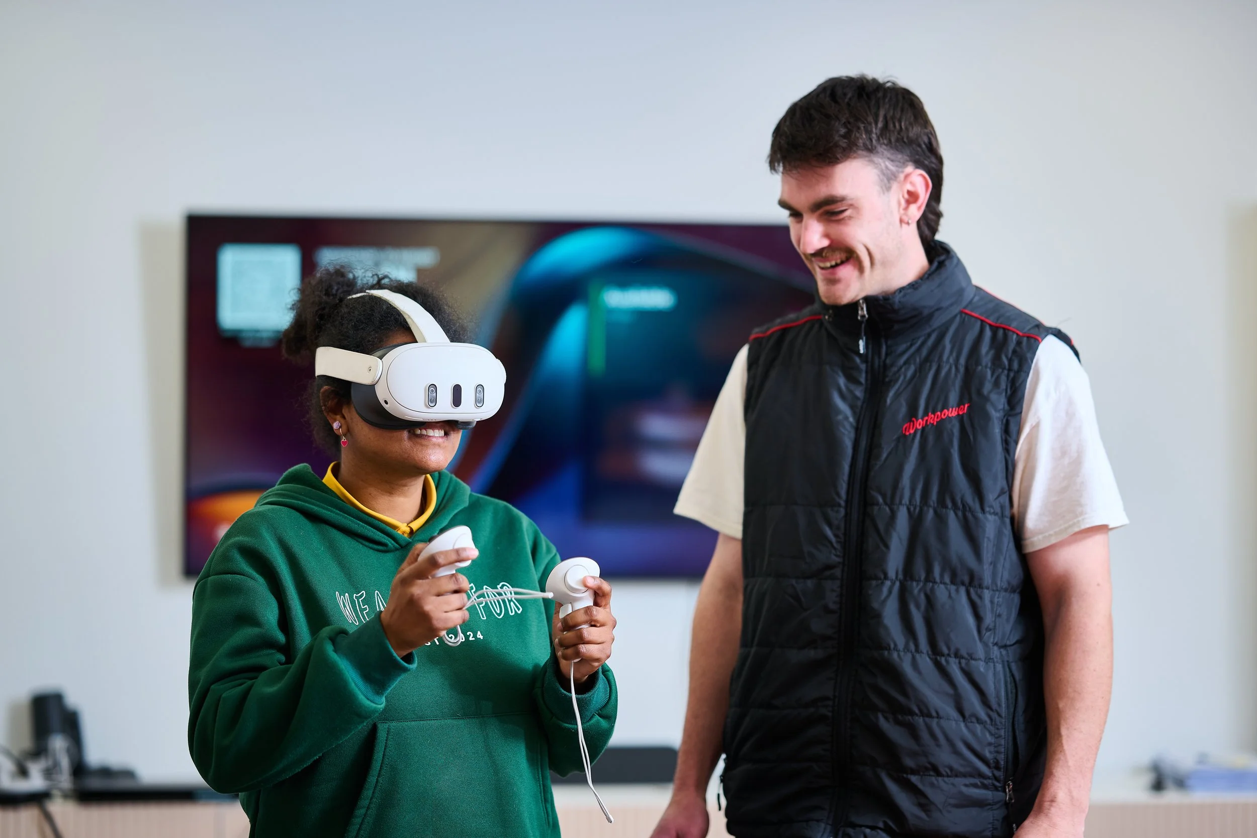 A woman uses a VR headset and controllers, exploring disability services as a man stands beside her, smiling in a modern indoor setting.