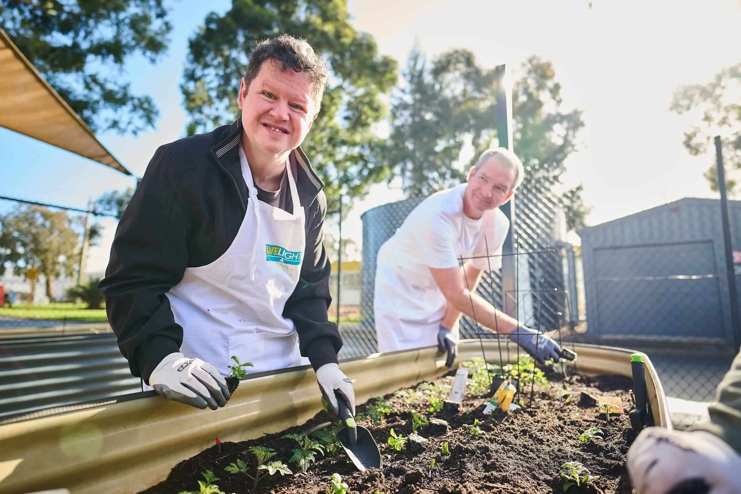 Two men wearing aprons and gloves, supported by Bunbury Disability Services, garden together and plant seedlings on a sunny day.