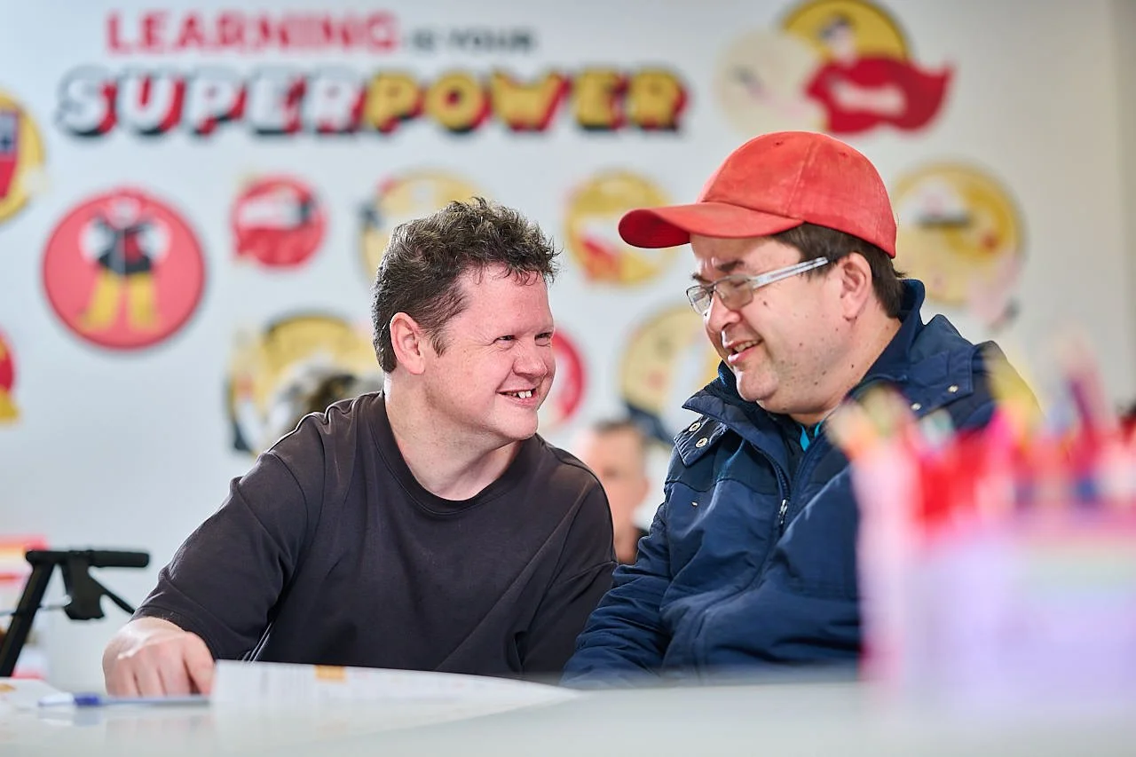 Two men smiling at a table, with colorful “Superpower” posters behind them, celebrating disability services and inclusion.