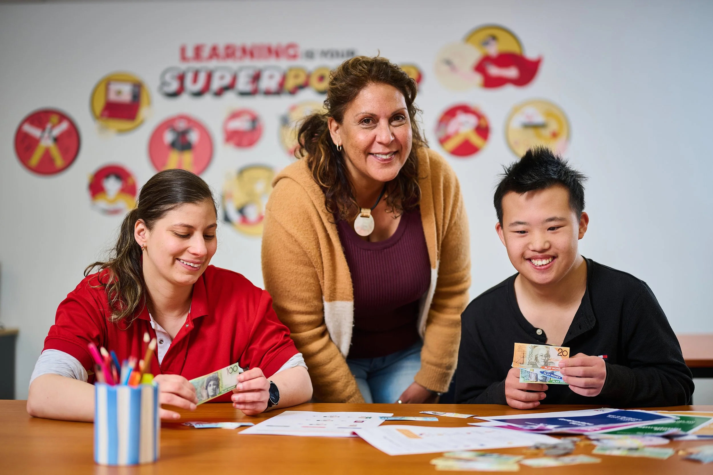 A teacher and two students sit at a table, smiling with money. Educational posters and disability services info are on the wall behind them.