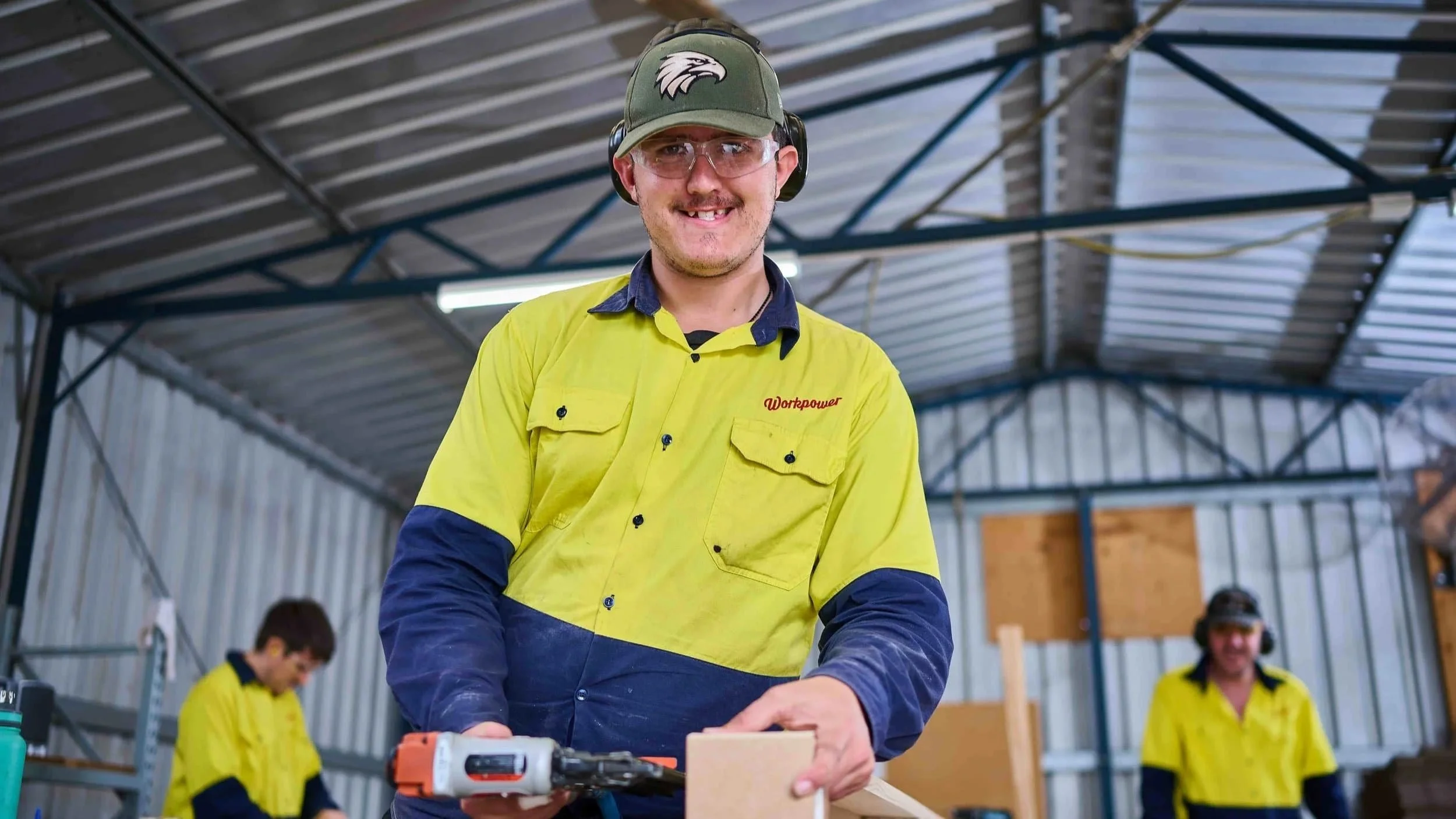 A smiling worker in a yellow safety shirt uses a power tool at Bunbury Disability Services, with others working in the background.