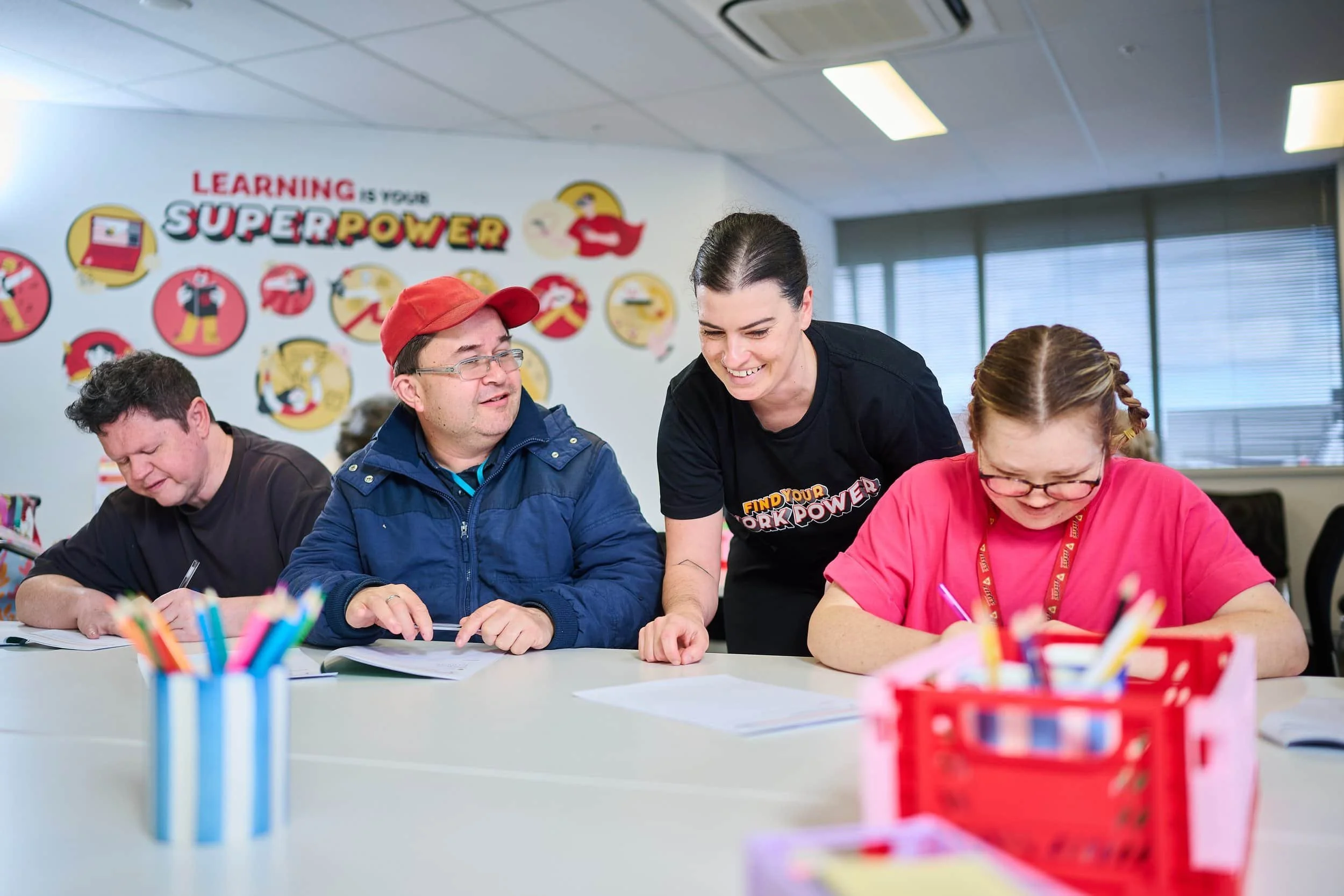 A teacher from Bunbury Disability Services assists three smiling students in a colorful classroom with a Superpower learning poster.