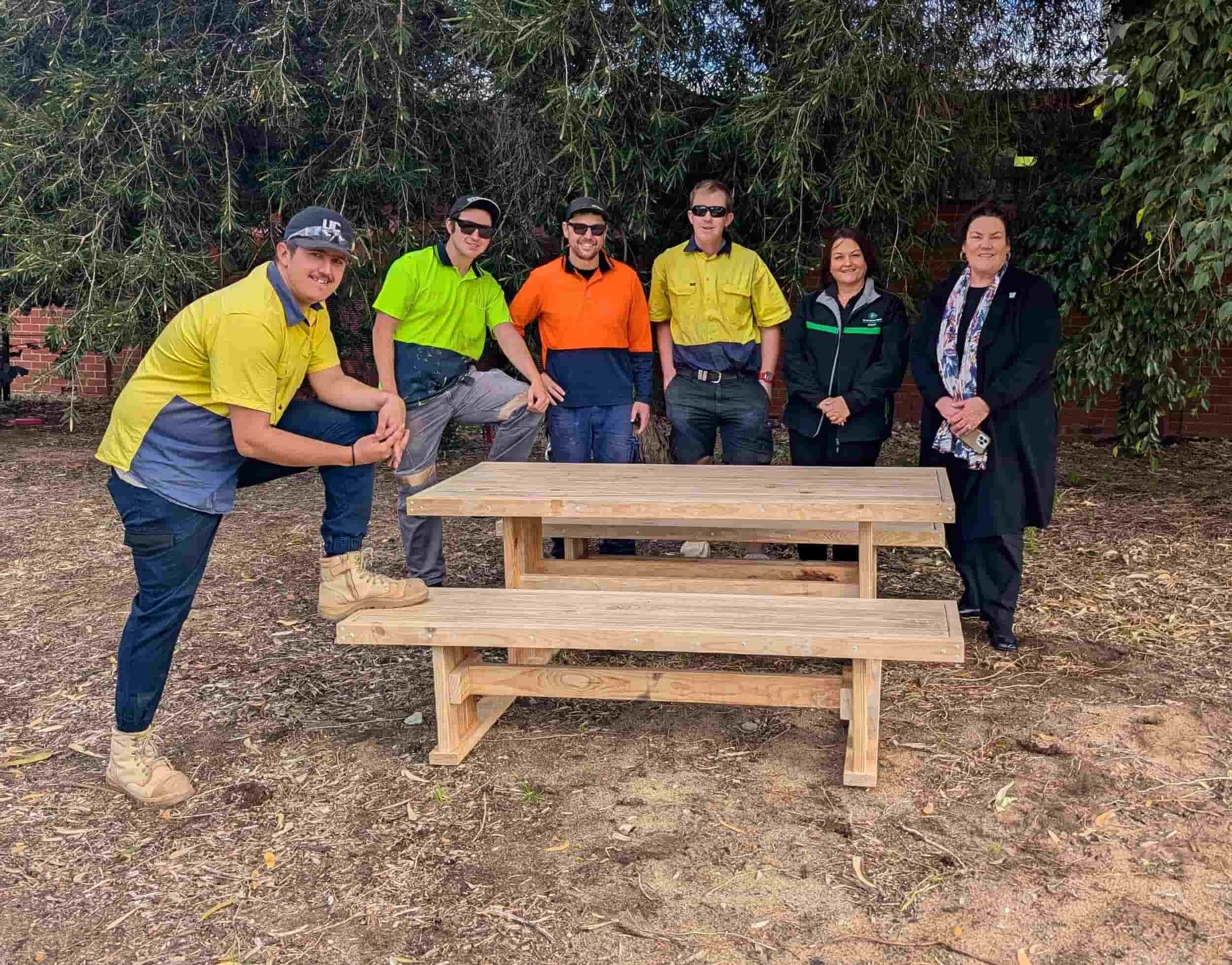 Six people smile behind a wooden bench outdoors, under a tree, celebrating community and disability services.