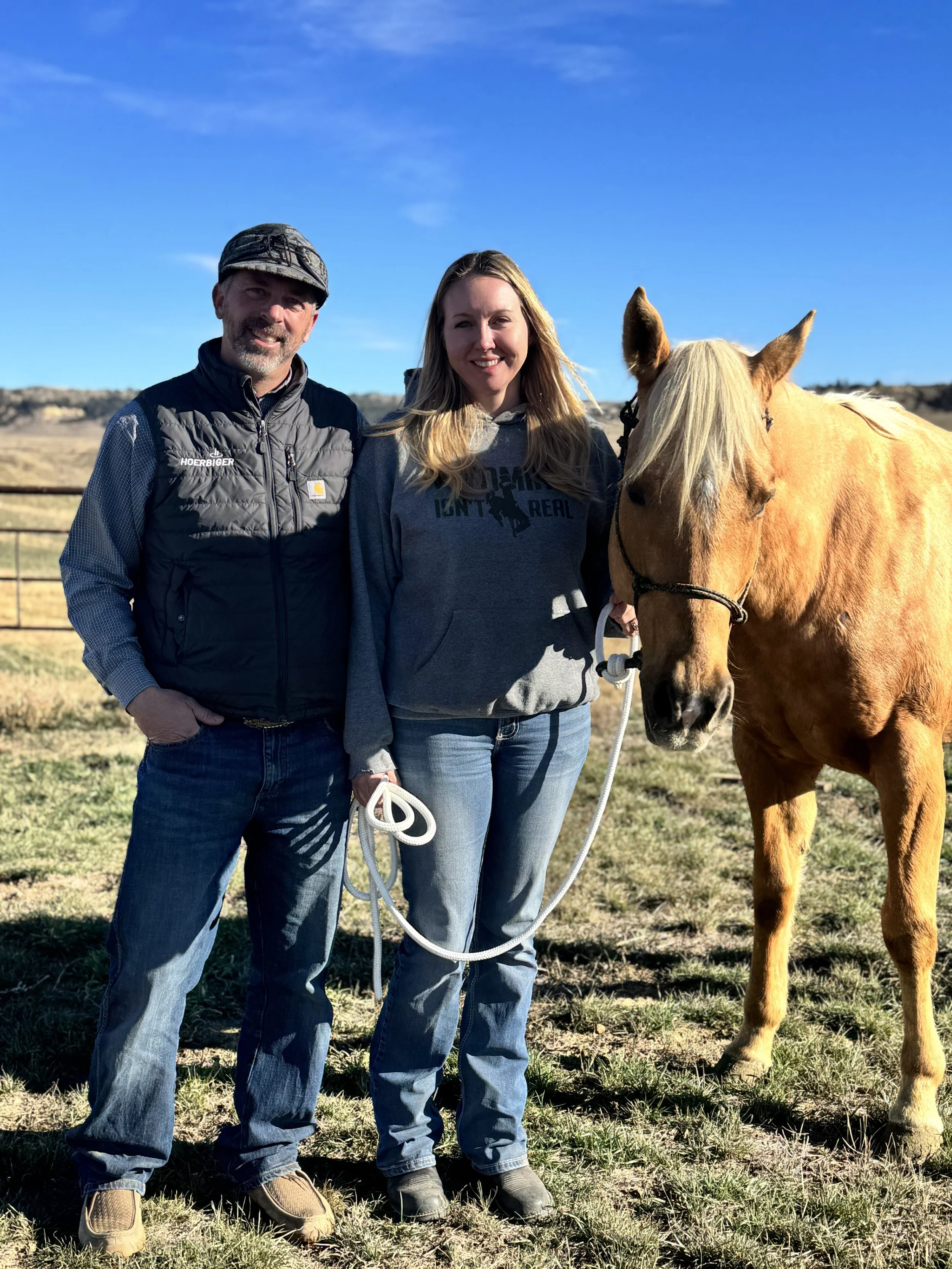 A man and woman standing outdoors in a pasture, smiling at the camera with a light brown horse next to them. The man is wearing a baseball cap, black vest, and jeans. The woman has long blonde hair, is dressed in a gray hoodie and jeans, and is holding a white rope attached to the horse’s halter. The background features a fence, open fields, and a clear blue sky.
